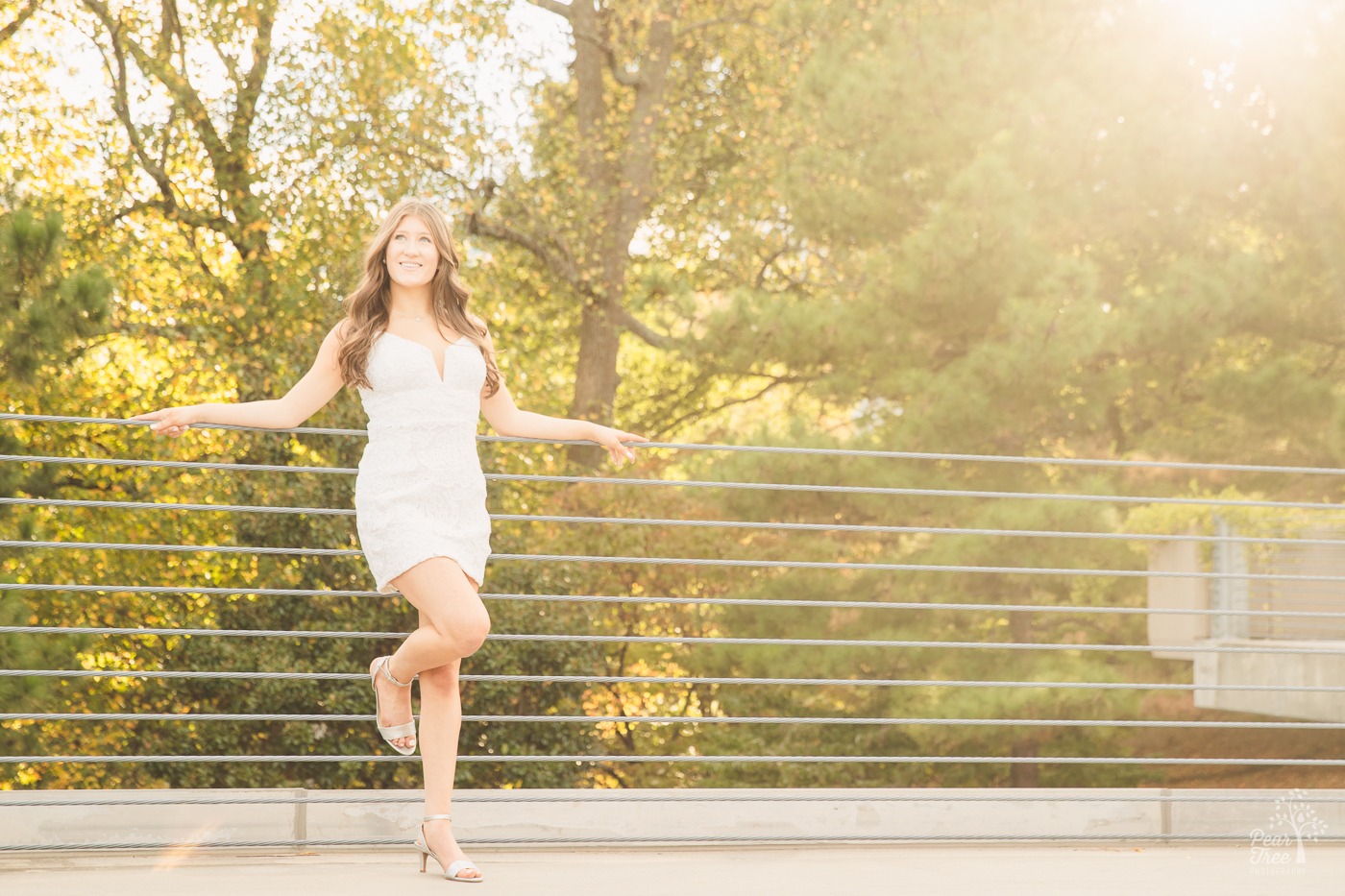High school senior girl leaning against wire railings and smiling in a white lacy dress with the sun shining through trees behind her