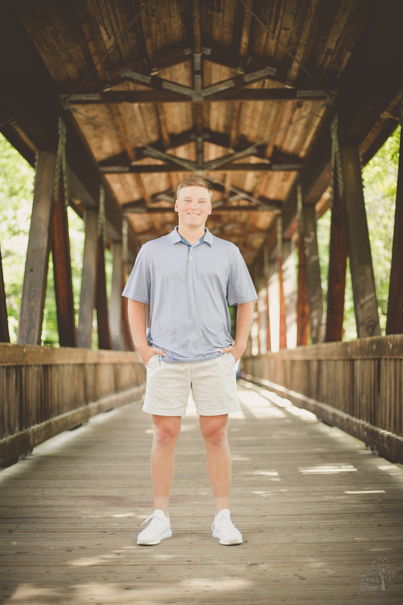 High school senior boy standing under covered bridge and hands in pockets while smiling