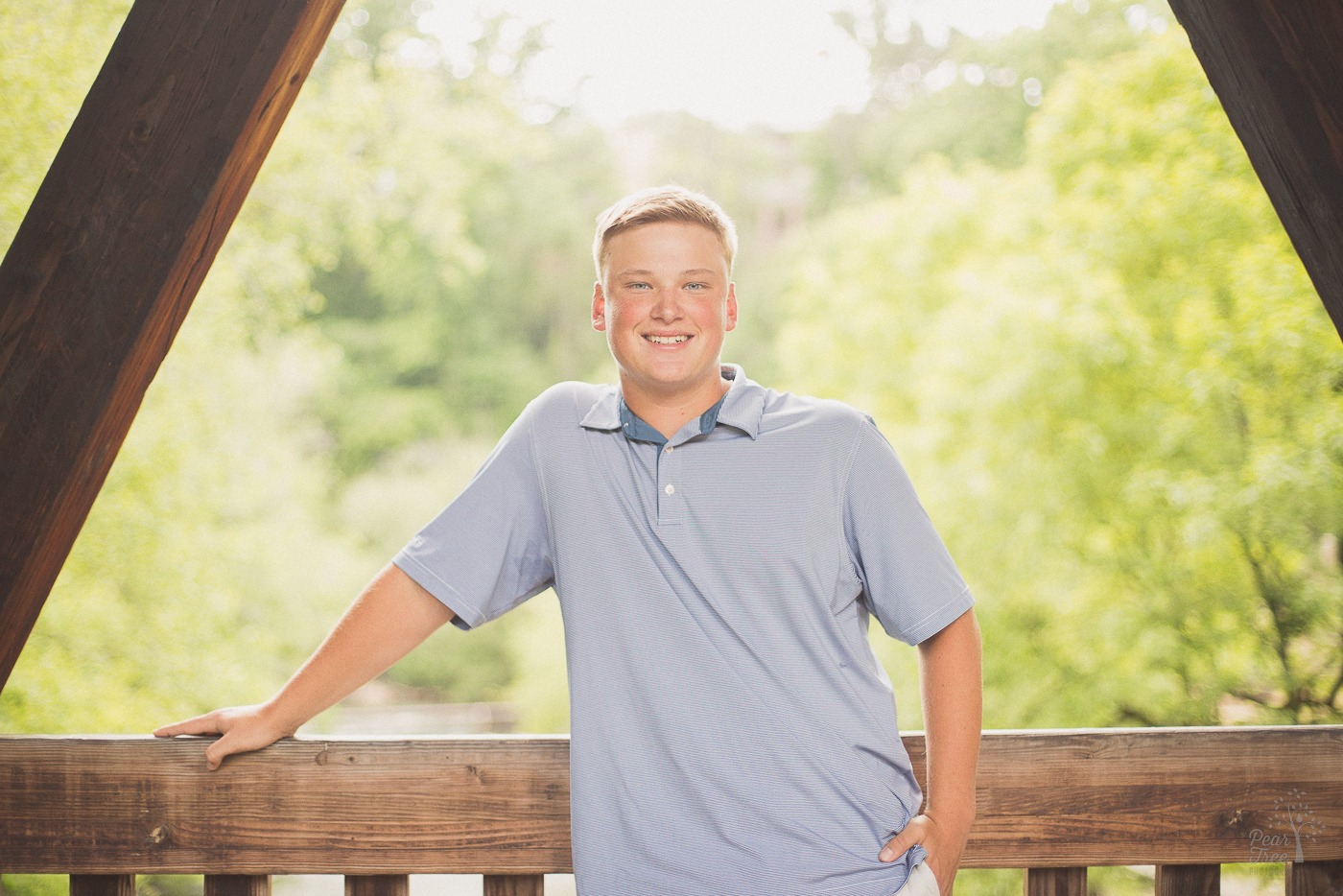 Happy teenage boy with hand in pocket and leaning on a wooden railing with trees and sun behind him