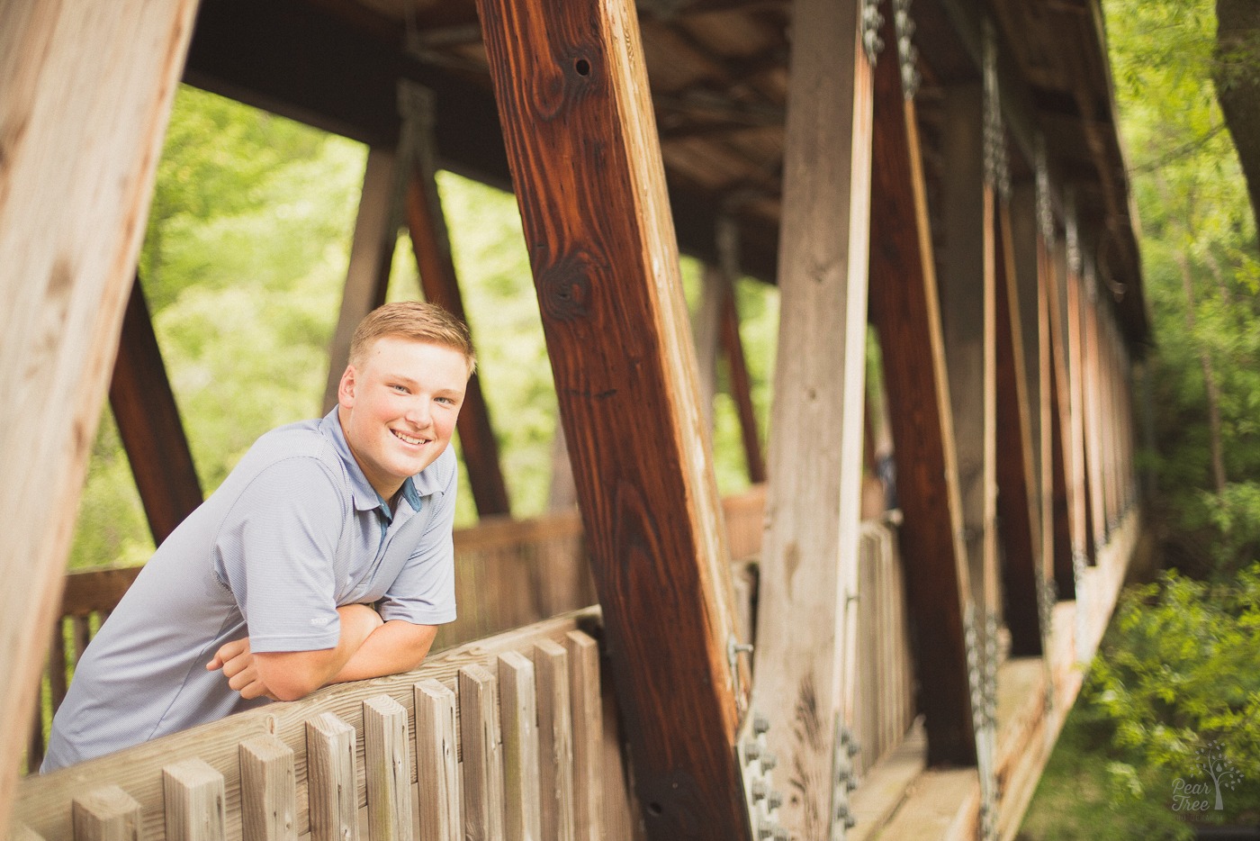Smiling high school senior boy leaning over railing underneath covered bridge and smiling at Roswell Mill