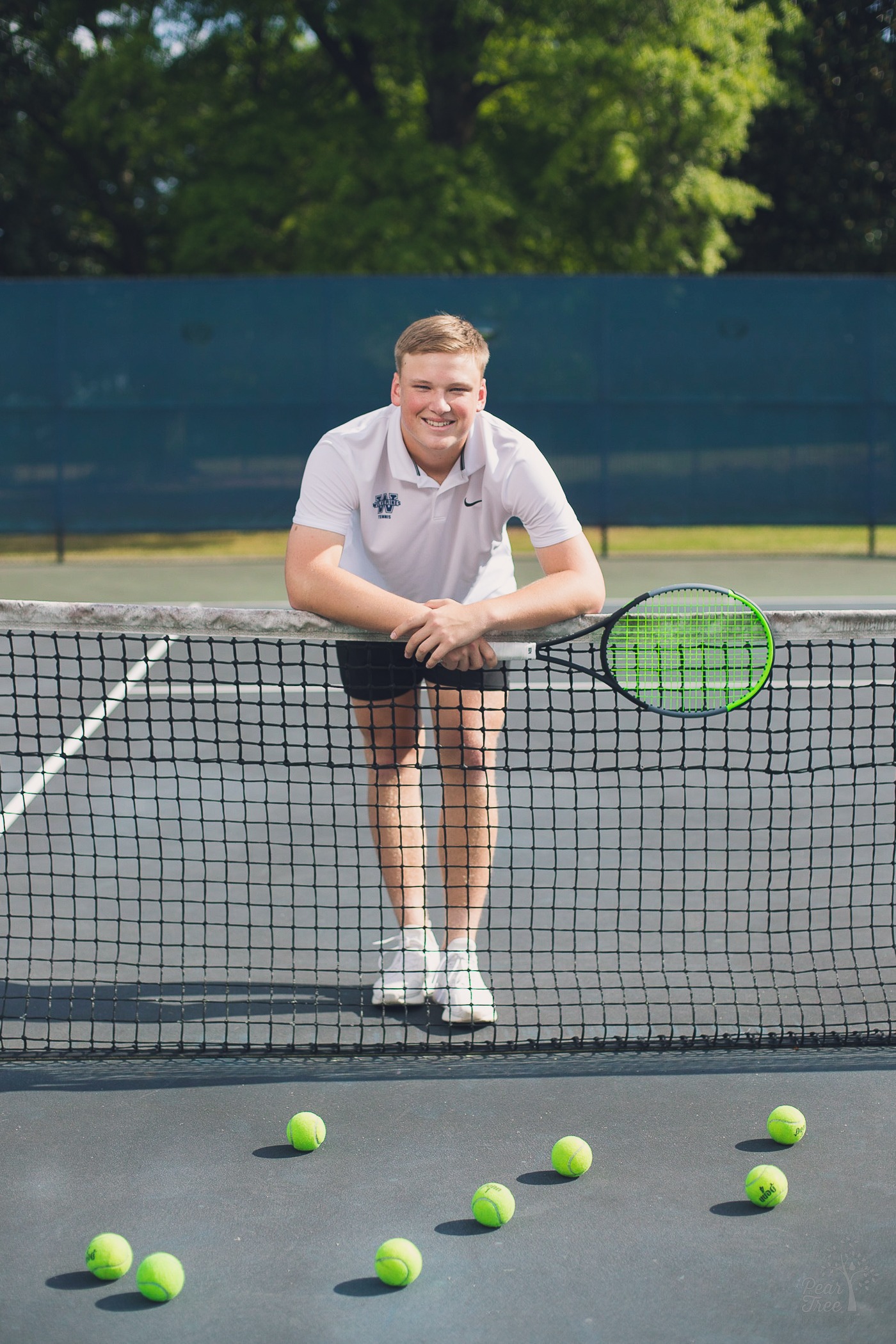 Smiling Woodstock High School Varsity Tennis senior leaning over tennis net with racquet in hand and tennis balls on the court in front of him