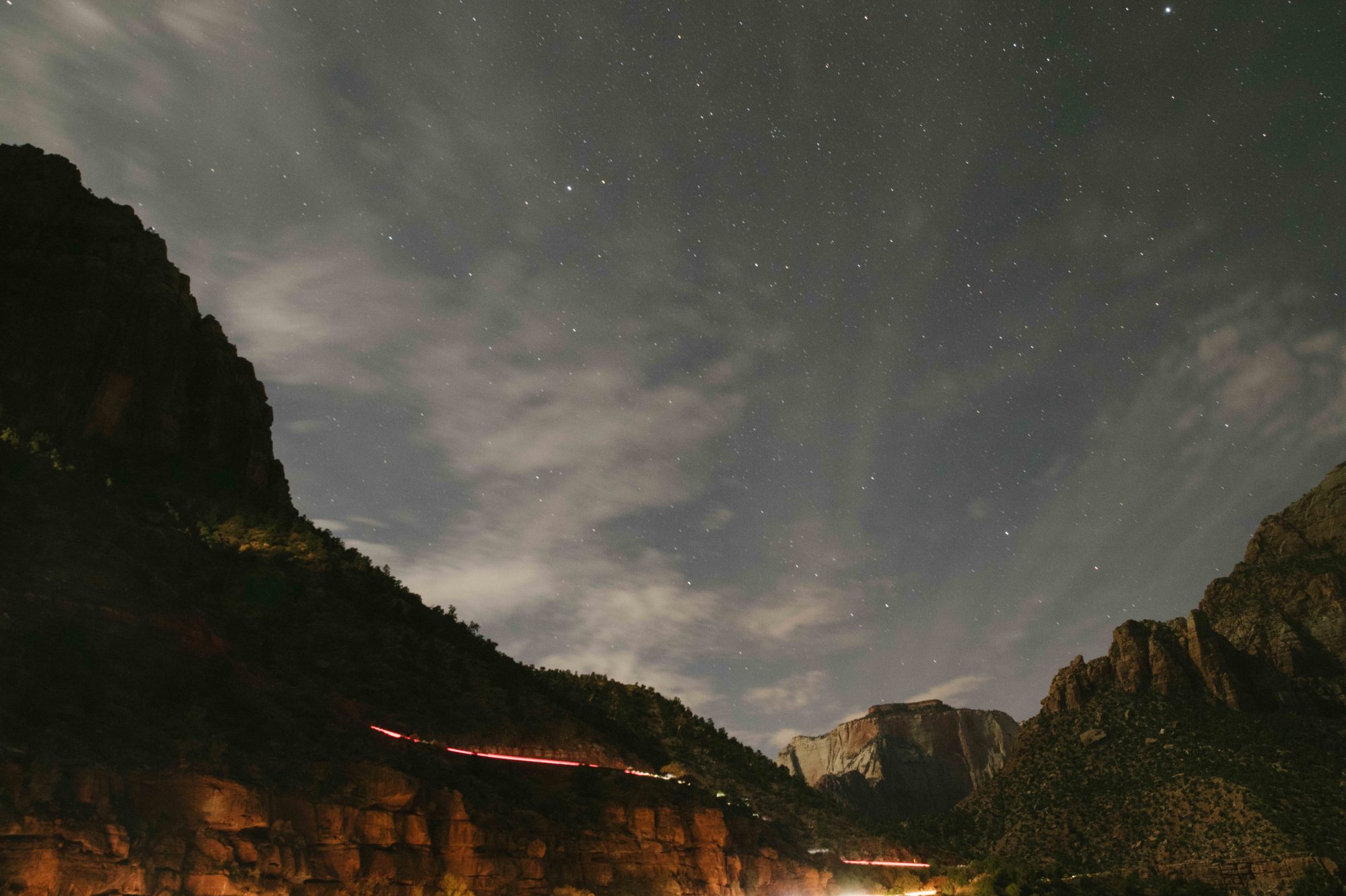 Night photograph of stars and clouds above Zion National Park with light trails from cars driving down the mountain road