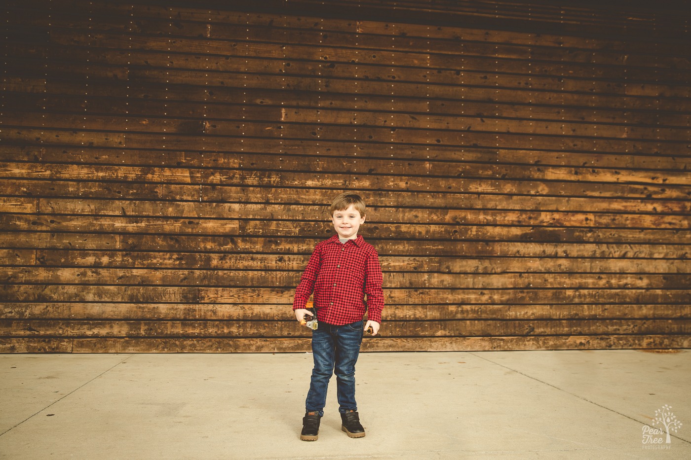 Little boy holding his toy train and smiling in front of a wooden slat wall