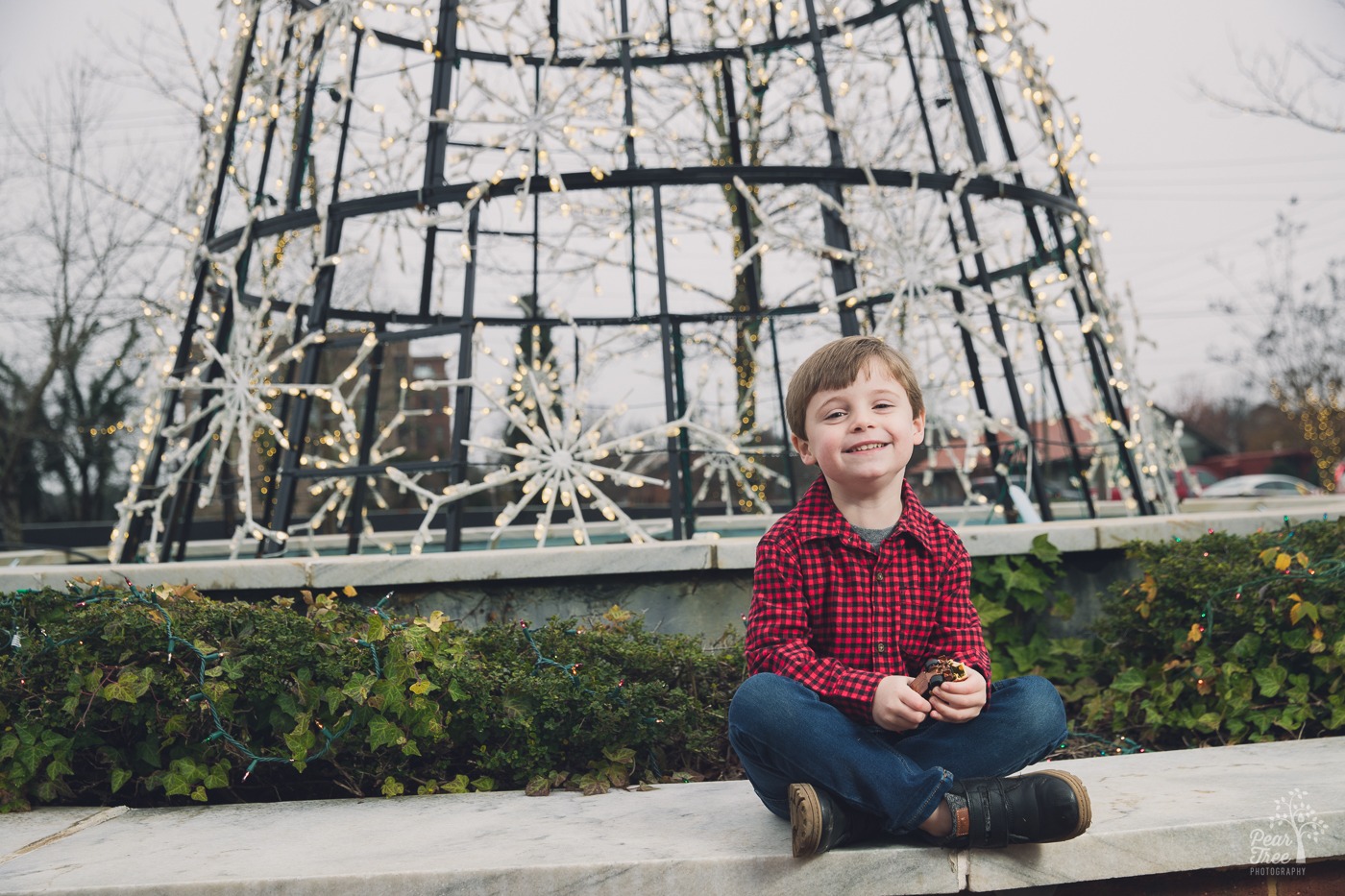 Little boy sitting criss cross applesauce in front of a Christmas tree made out of snowflake shaped lights in Downtown Woodstock