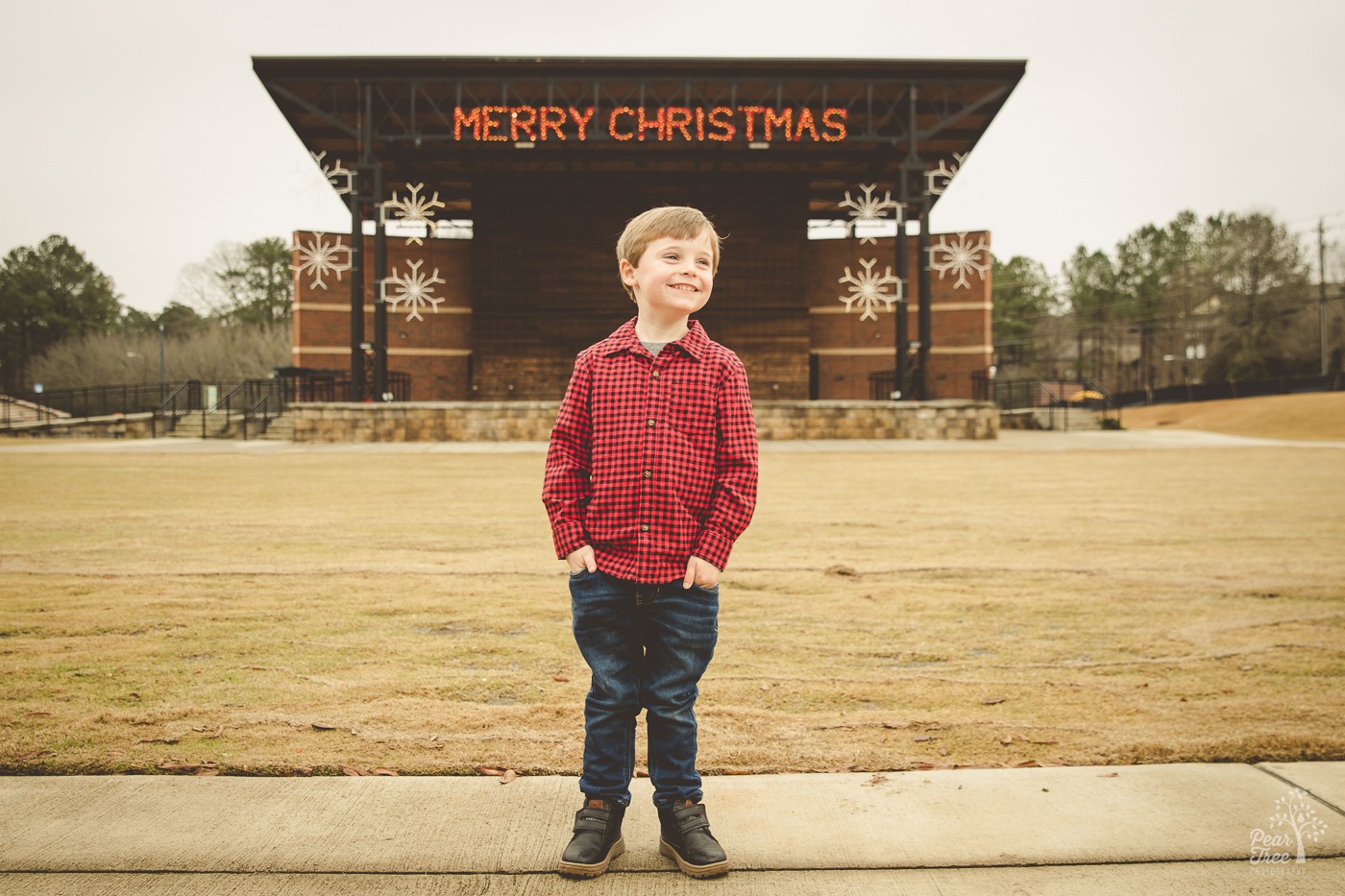 Cute little boy smiling with his hands in pockets with downtown Woodstock amphitheatre behind him. Merry Christmas and snowflake lights are lit up