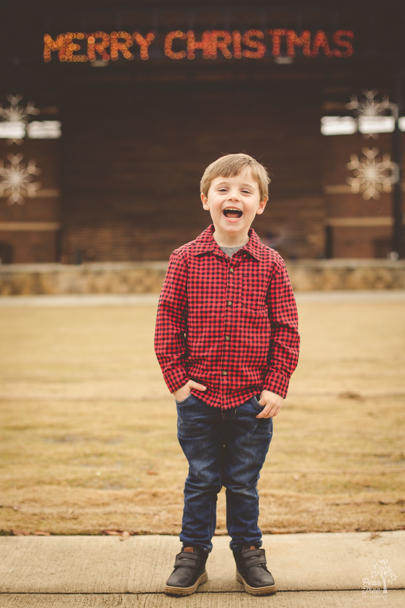 Little boy laughing big with a Merry Christmas sign lit up behind him during his four year old photoshoot in downtown Woodstock