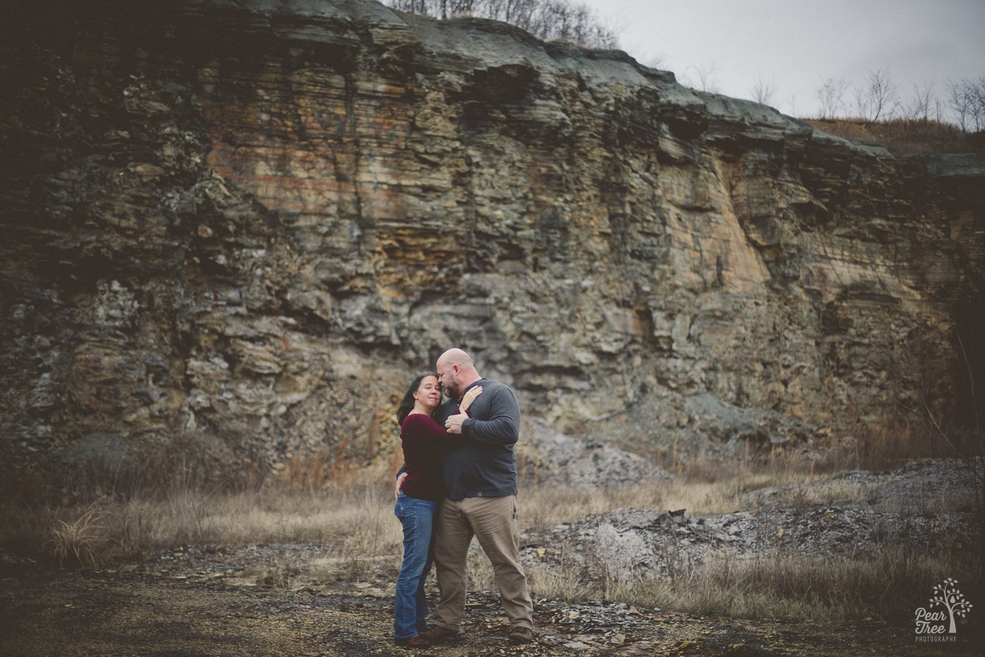 A tall man holding his girlfriend close while he looks at her and she looks at the camera. They're standing in front of a tall shale wall