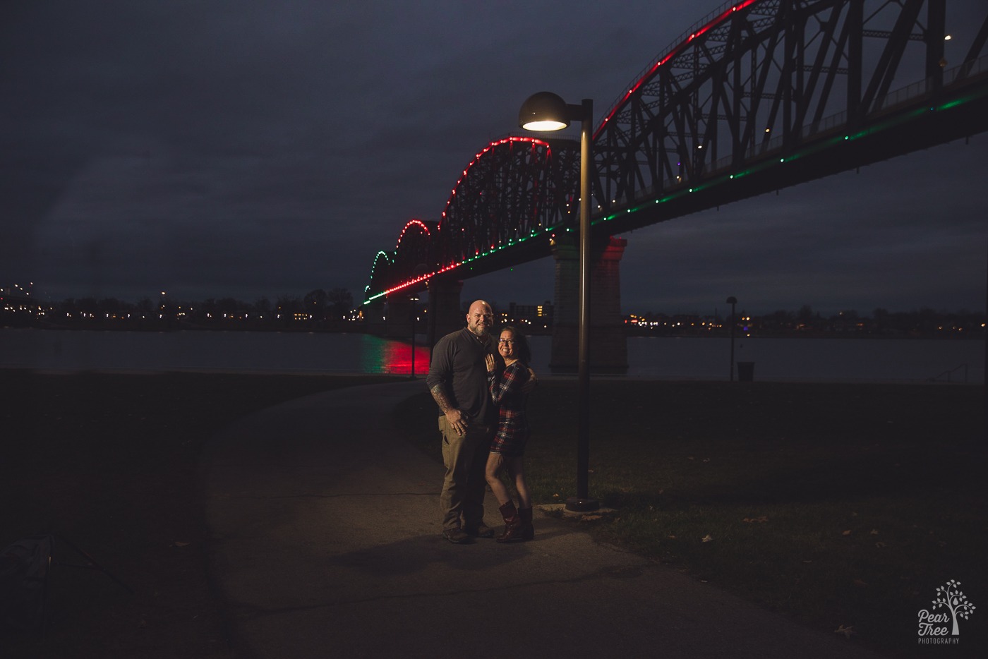 Smiling couple looking at the camera while standing under a street light beneath the Big Four Bridge in Louisville