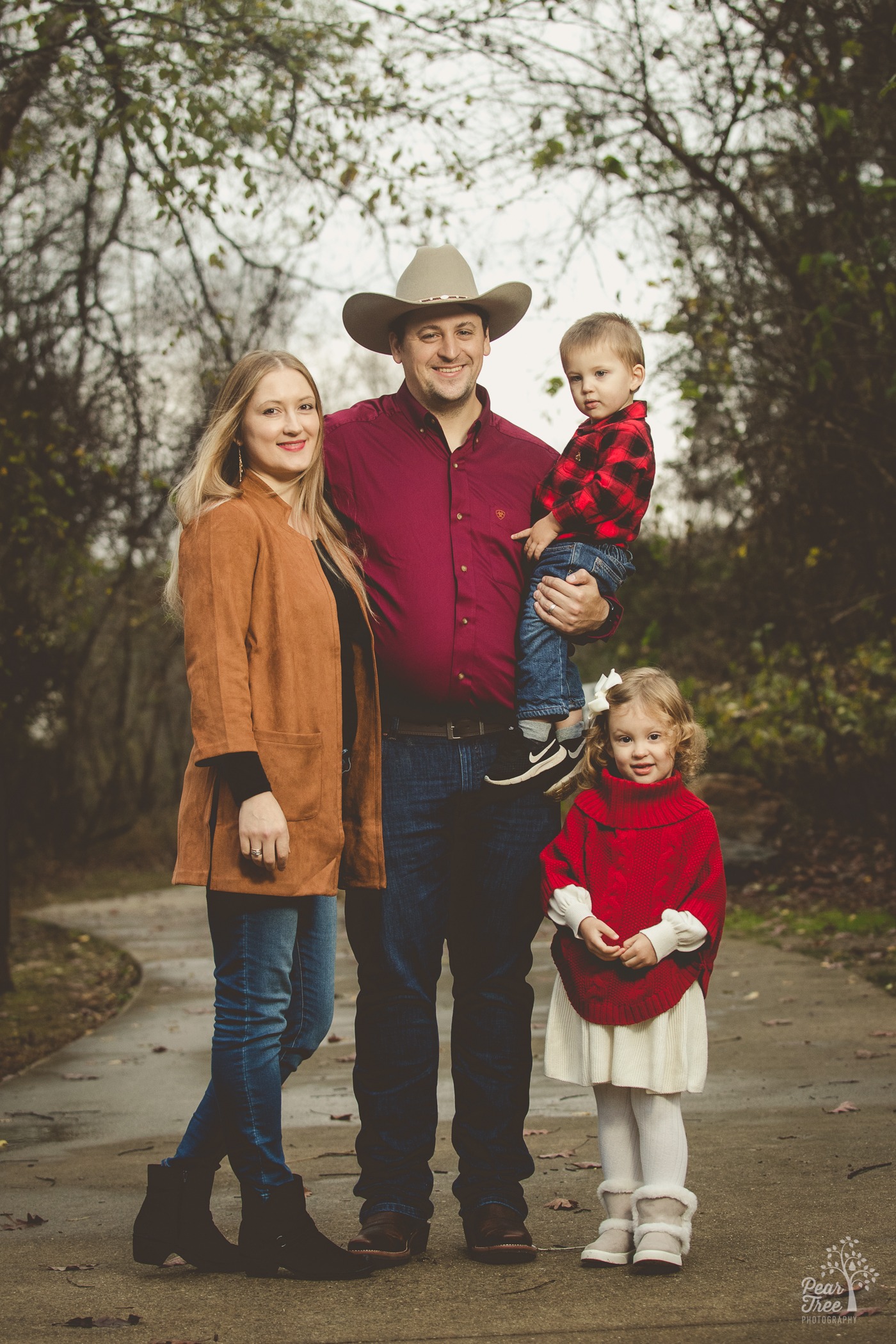 A cute family with dad wearing a cowboy hate and holding his son while standing with his wife and daughter at a park in Woodstock