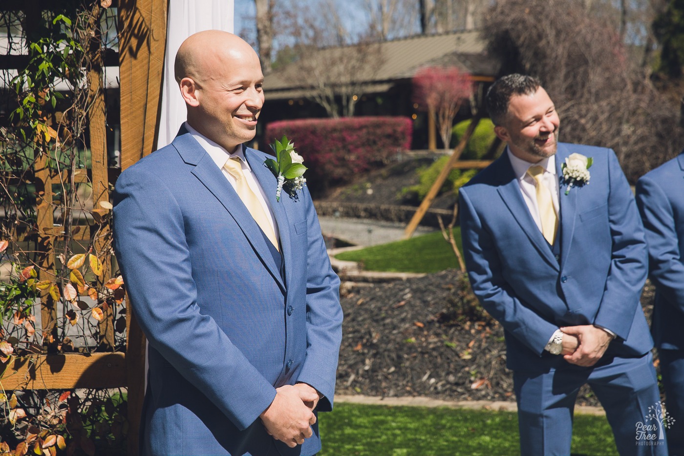 Groom smiling while watching his bride walk down the aisle at Rocky's Lake Estate while his groomsman leans in for a better look