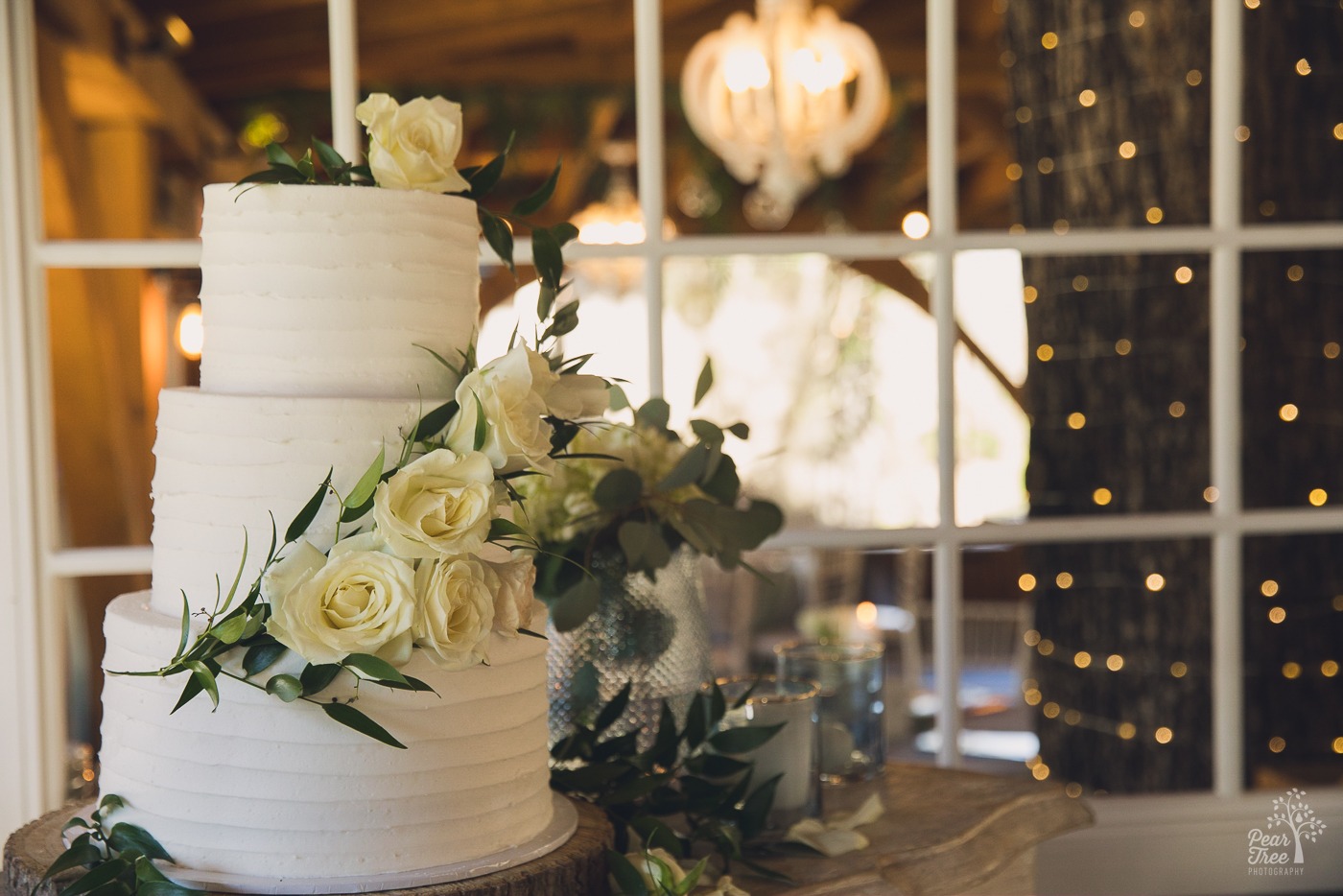 Three layer wedding cake with white roses wrapped around it and twinkle lights in the background