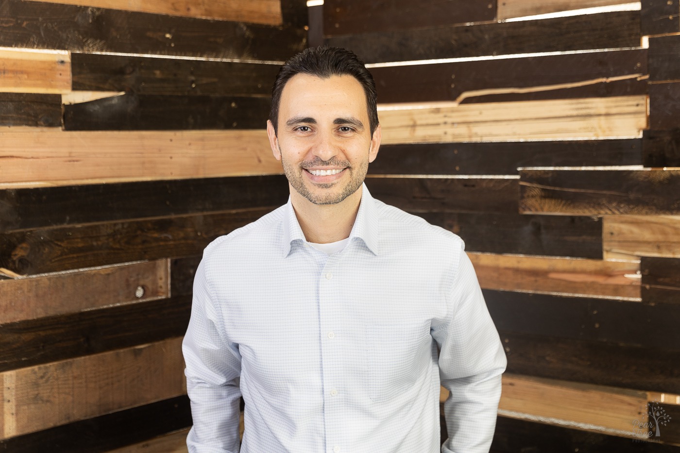 Smiling man in a dress shirt standing in front of a pallet wall with light shining through the cracks during professional headshots in a Canton studio