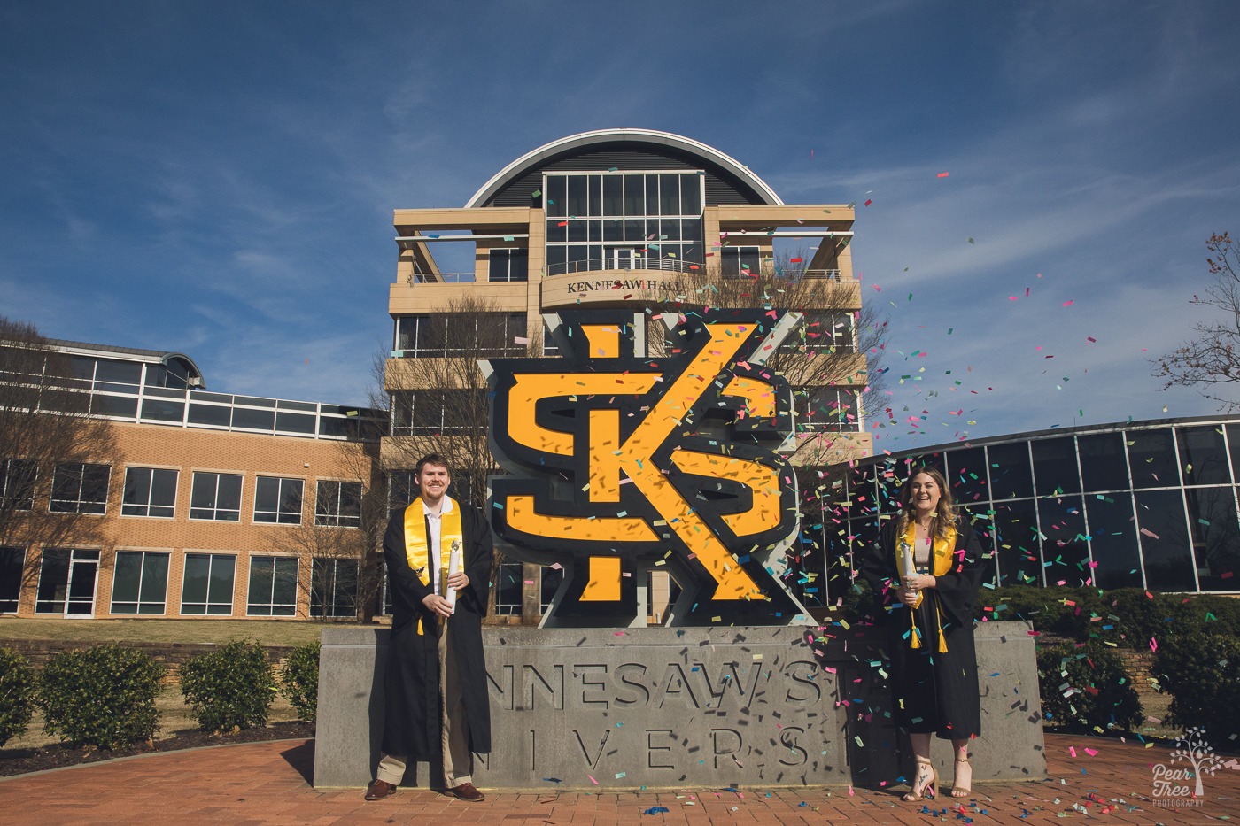 Two KSU graduation photos of two smiling graduates as their confetti falls to the ground in front of the KSU sign