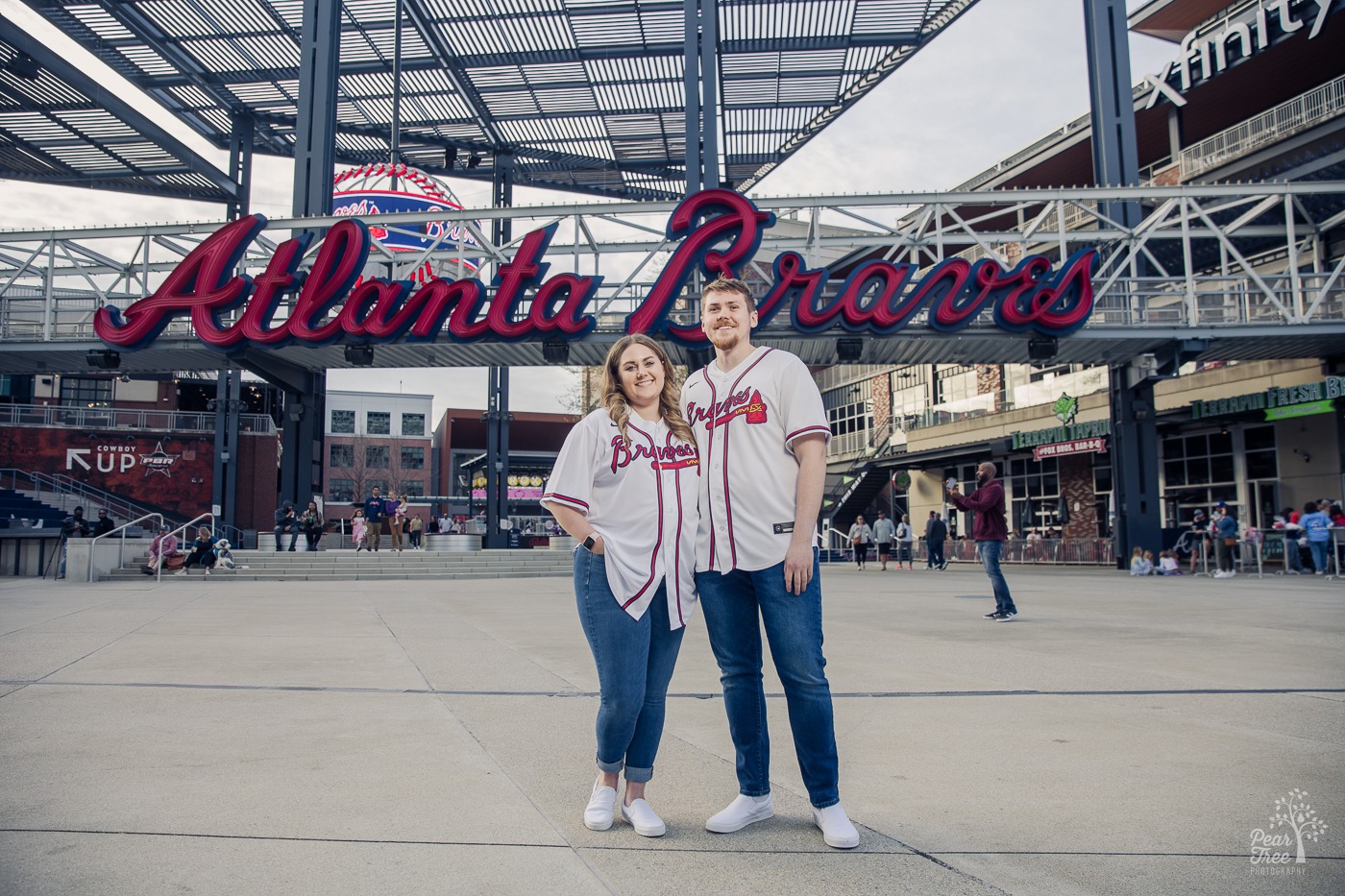 Smiling engaged couple wearing Braves jerseys and jeans in front of Atlanta Braves sign at the Battery