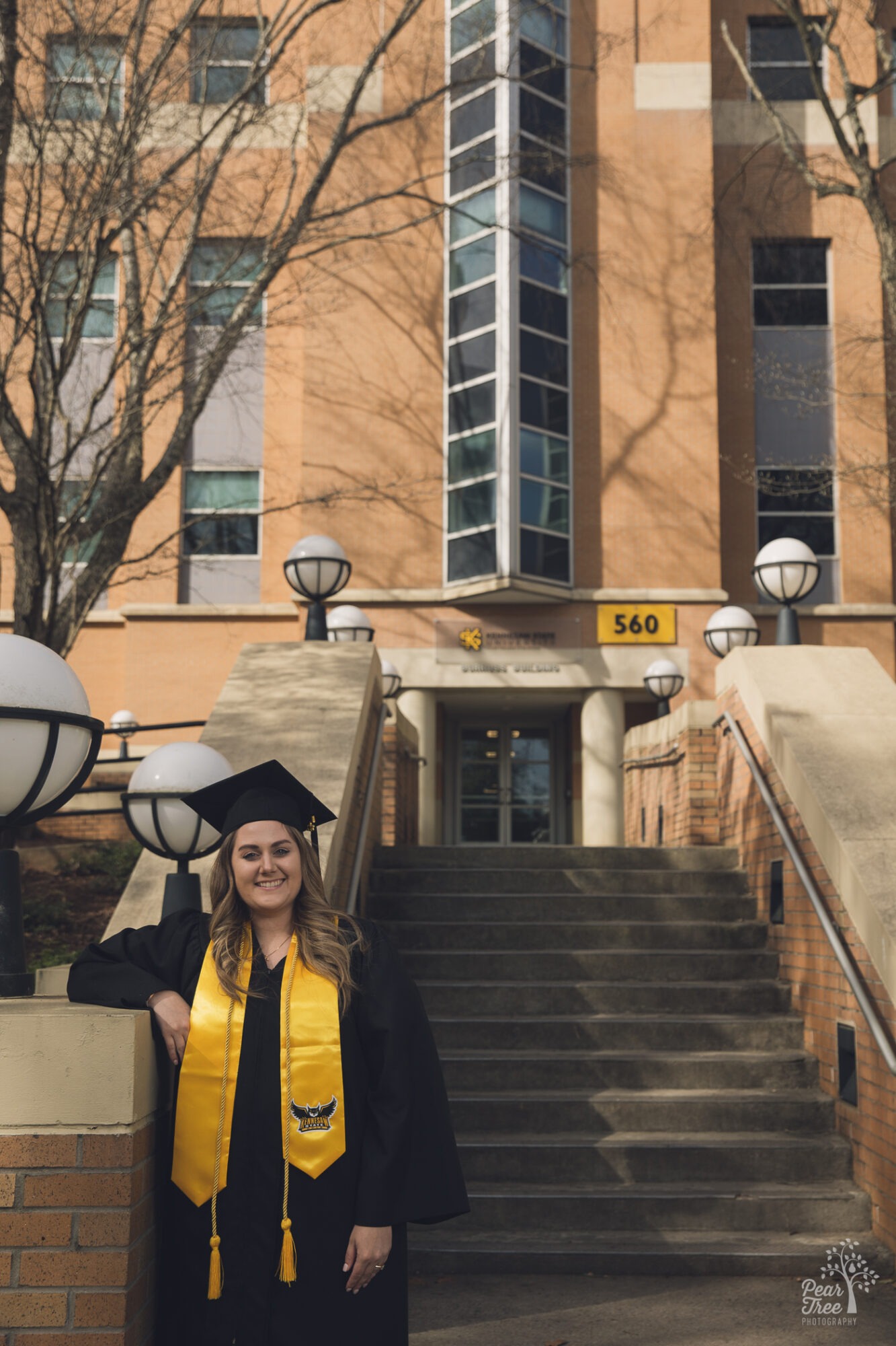 Smiling woman standing in cap and gown in front of Burruss business school for KSU graduation photos