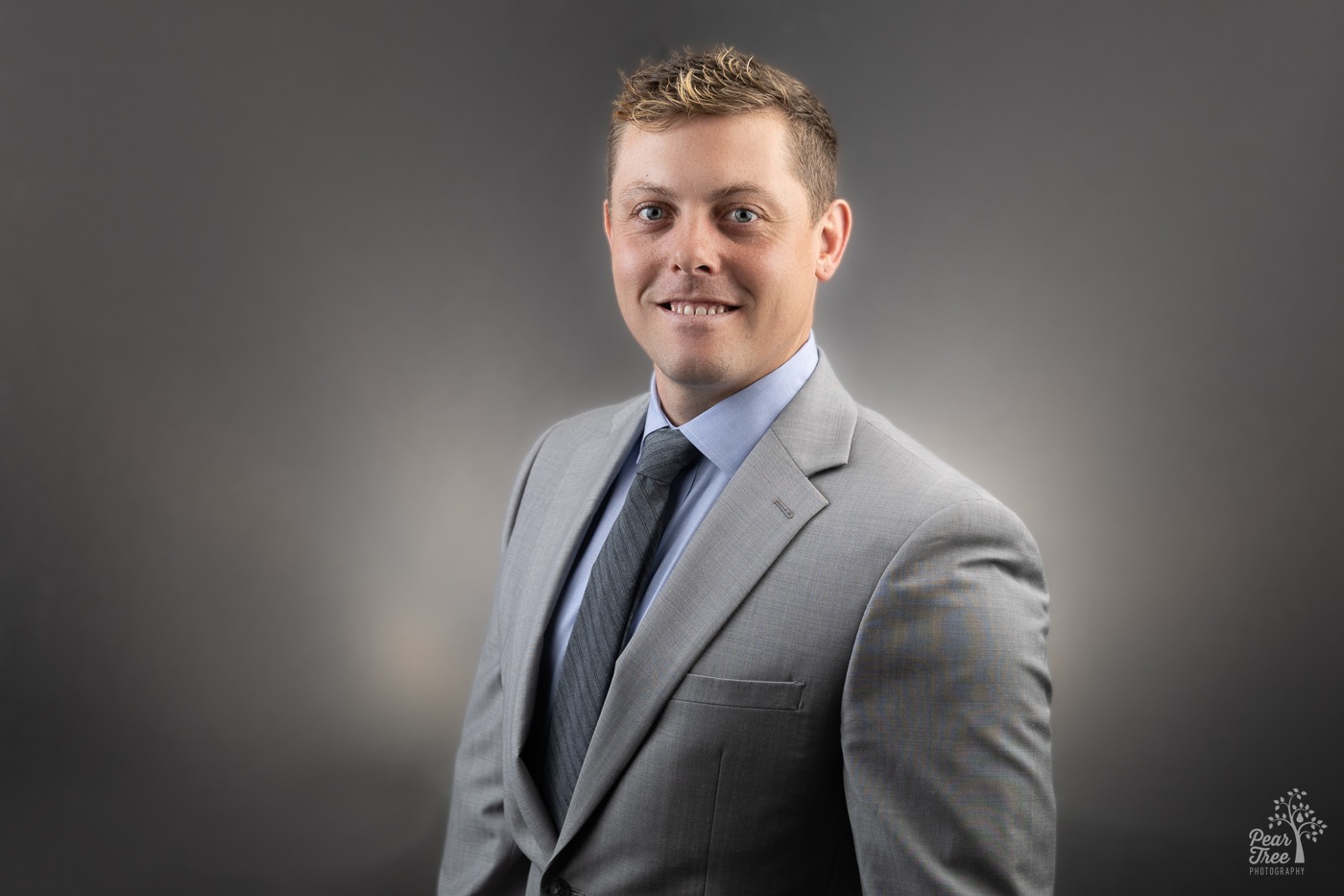 Smiling blond man in a grey suit and blue tie standing in front of a grey headshot background in Canton