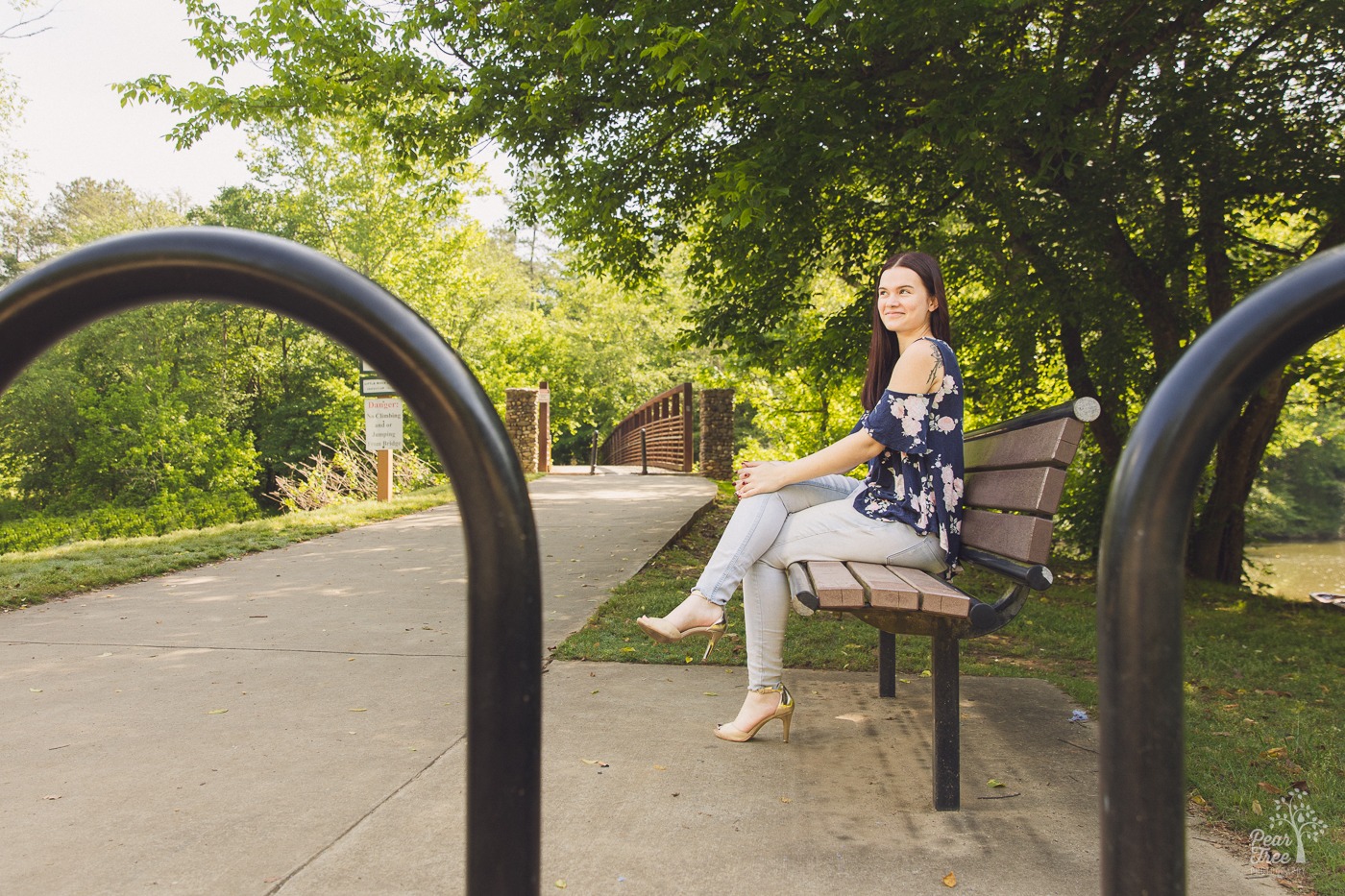 Looking through bike rack bars at a high school senior sitting on Olde Rope Mill Park bench with bridge in the background