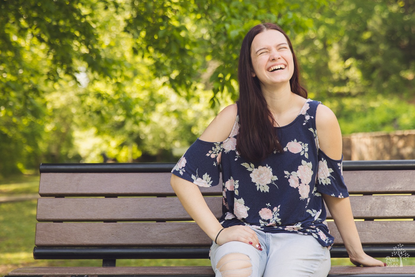 Laughing high school senior sitting on a park bench with trees in the background