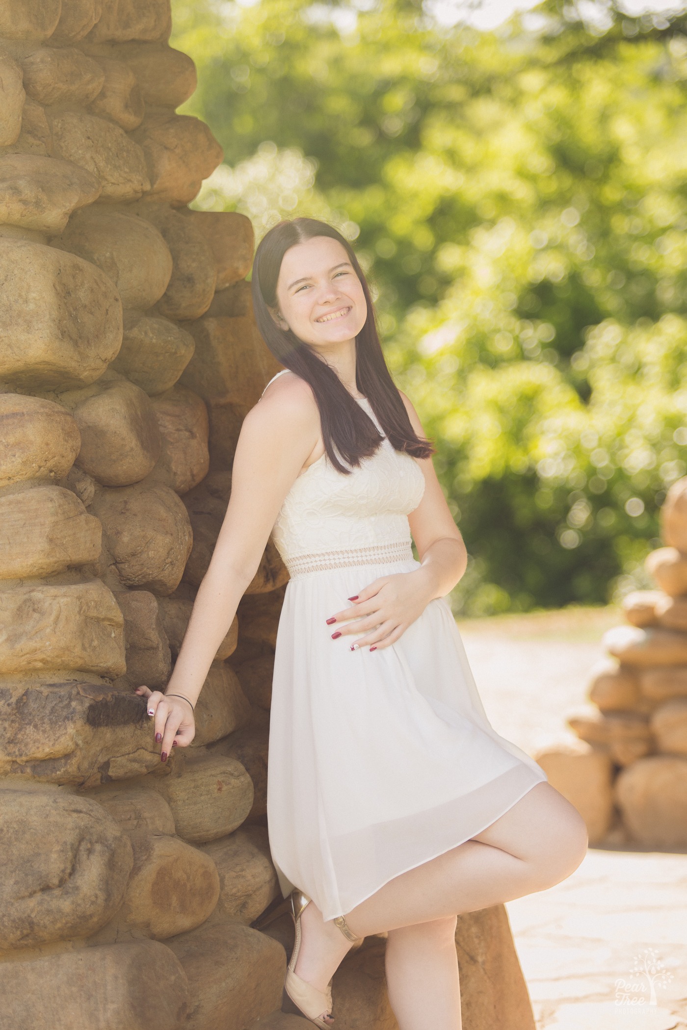 Beautiful high school graduate in white dress leaning against a stone wall