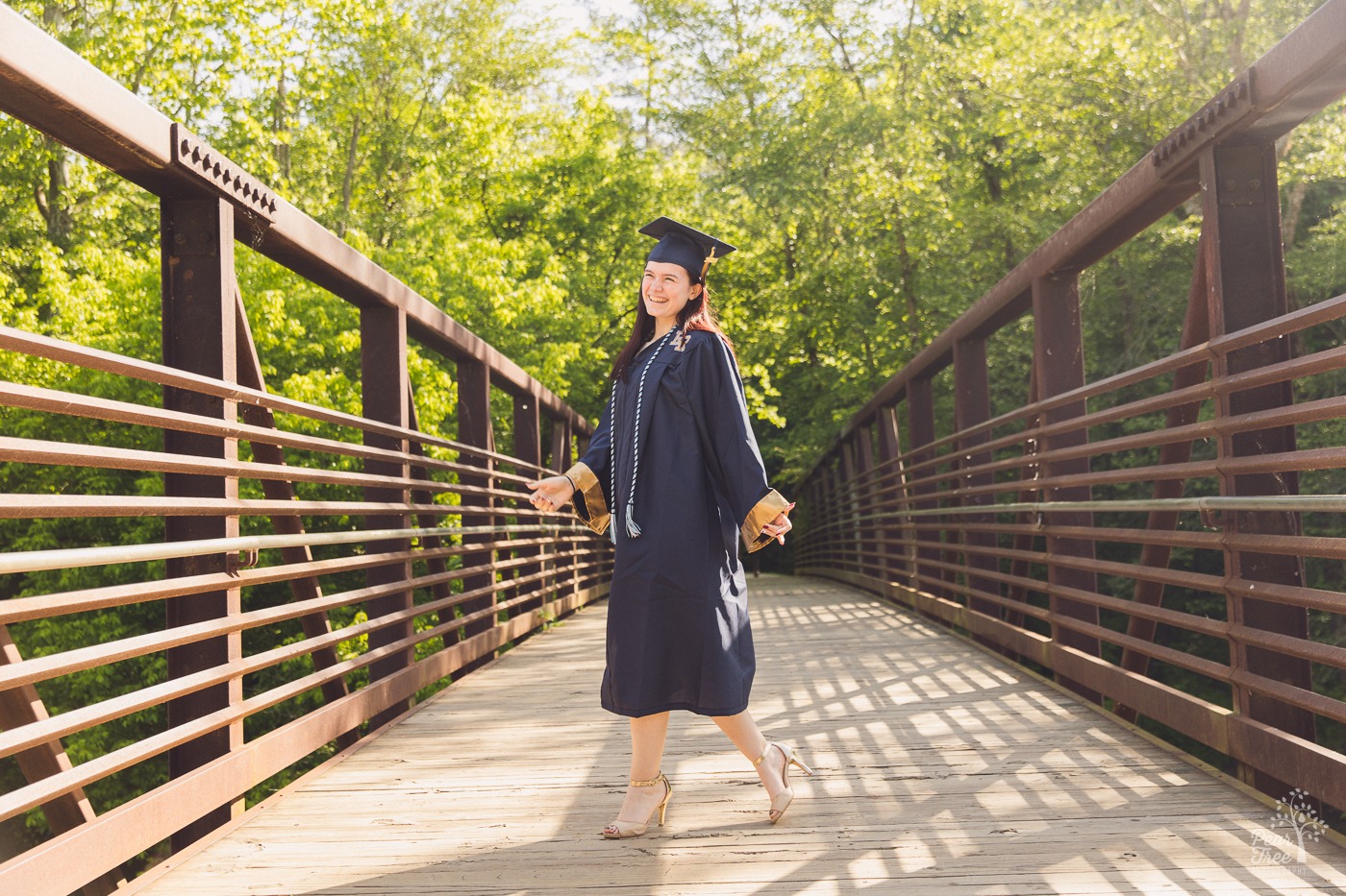 River Ridge High School senior in cap, gown, and high heels walking across Olde Rope Mill bridge