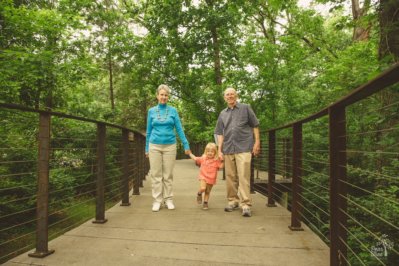 Grandparents smiling and hold their three year old grandson's hands as they walk over a bridge for multi-generational family photos
