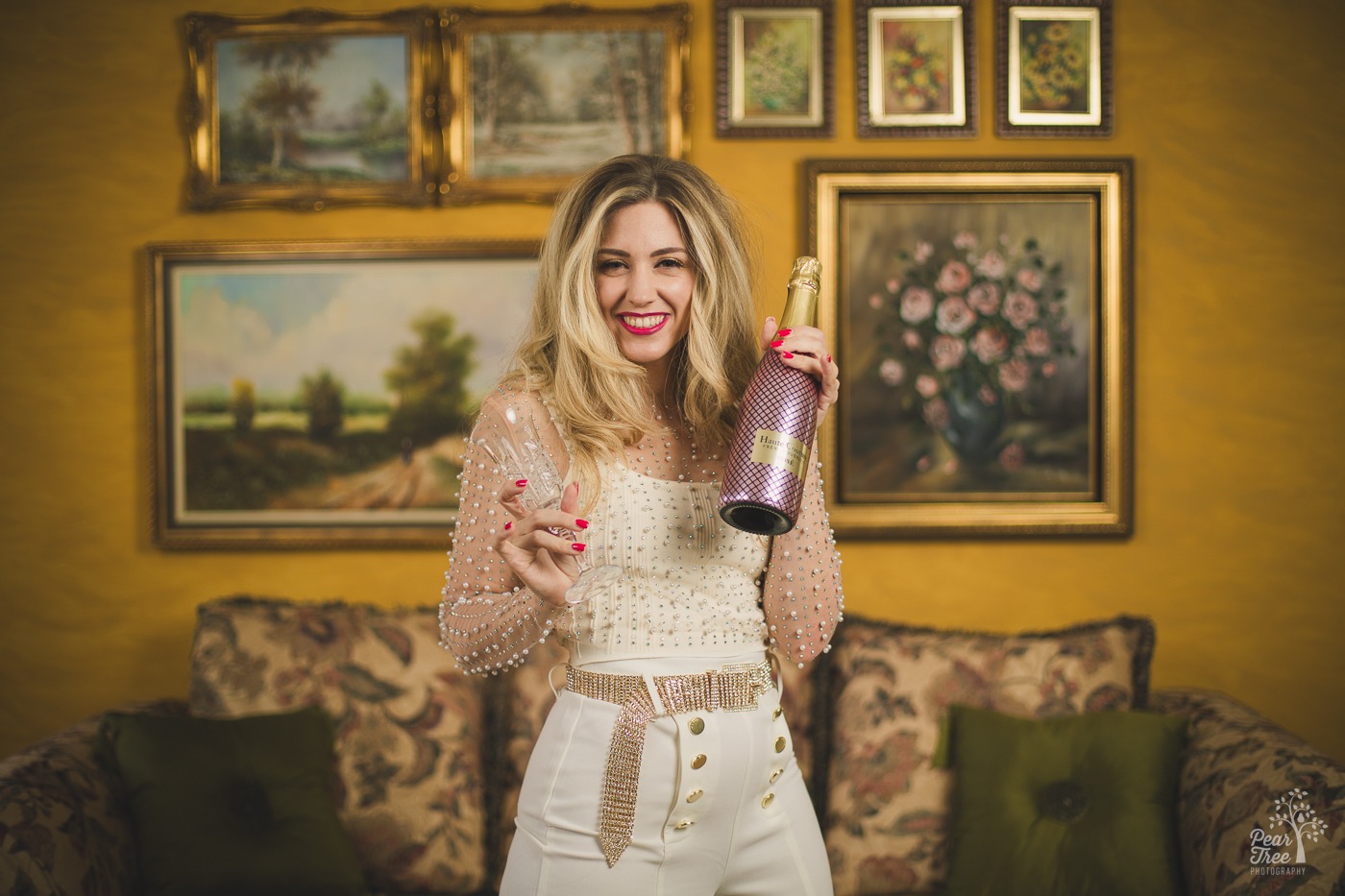 Alley Bei smiling while holding a bottle of champagne and a crystal glass with her mom's paintings hung on the wall behind her