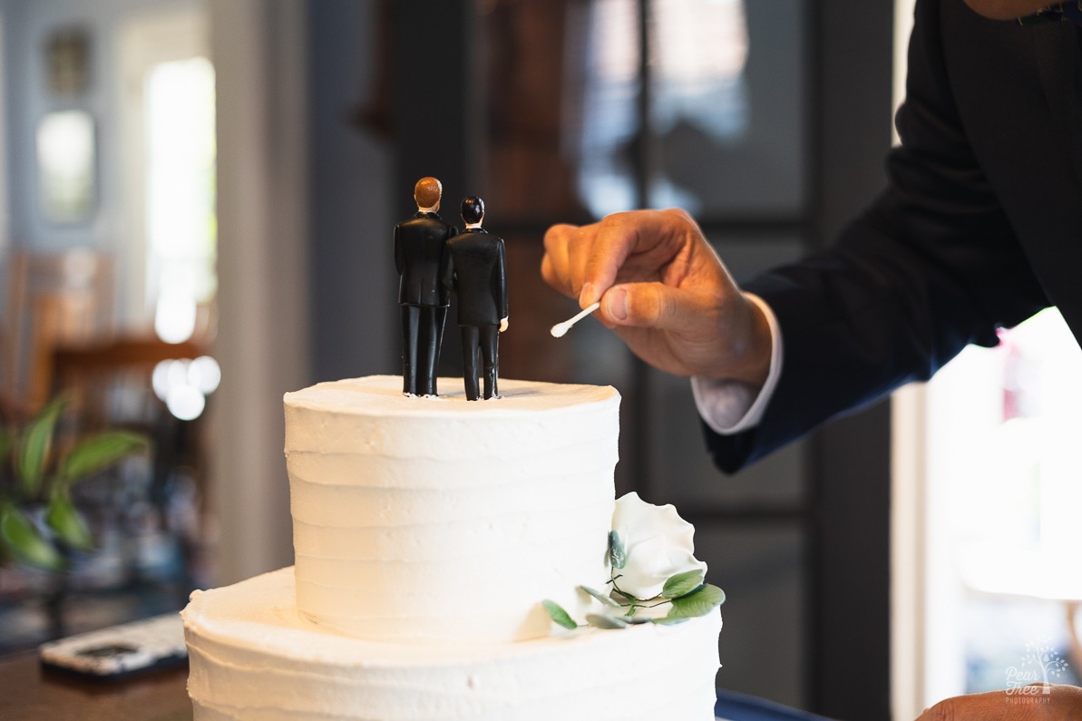 The arm and hand of a groom holding a q-tip to remove icing from groom figurines after placing them on top of the wedding cake