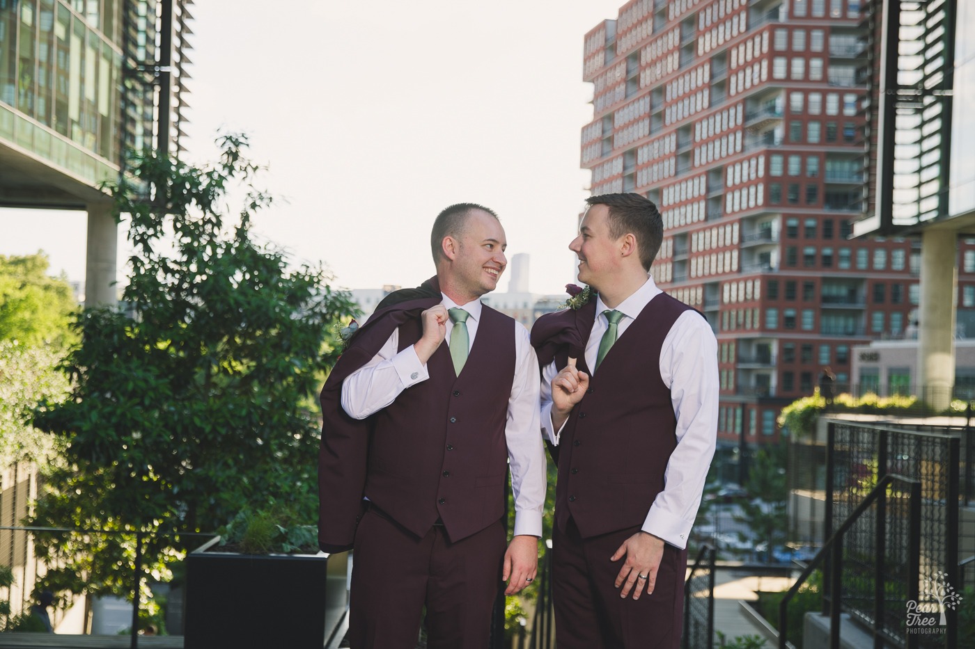 Two grooms smiling and standing casually holding their suit jackets over their shoulders