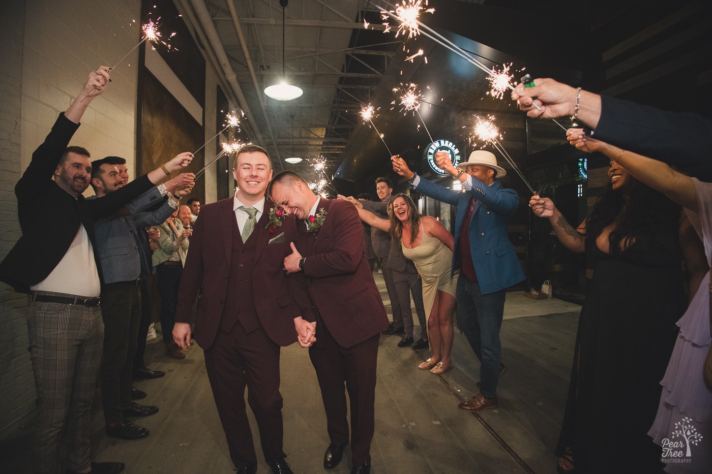 Two grooms holding hands and smiling as friends hold sparklers overhead while cheering