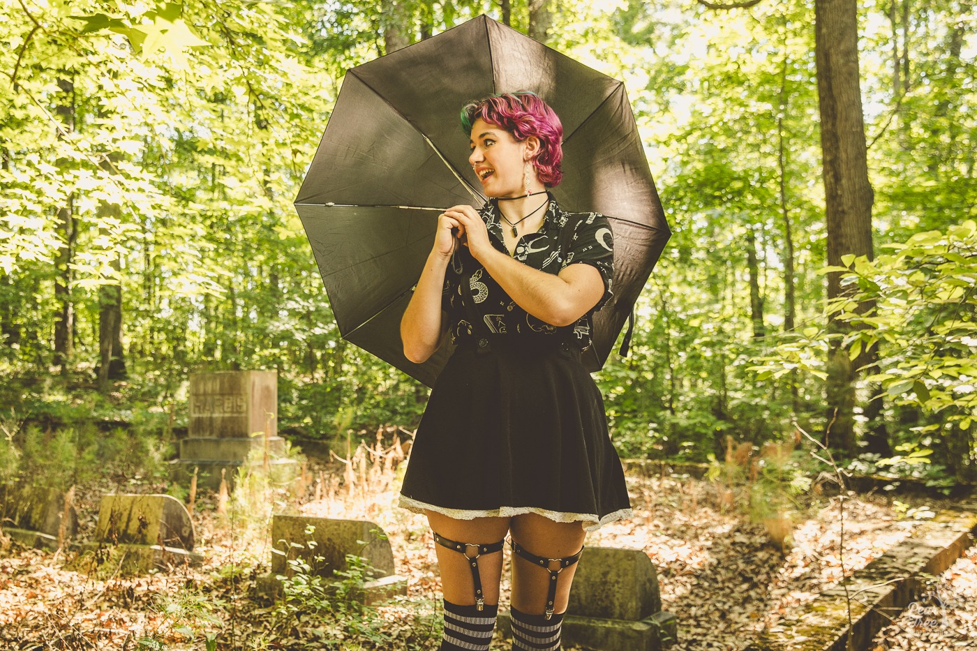 Teenager dressed in black holding a black umbrella and standing in front of an old cemetery