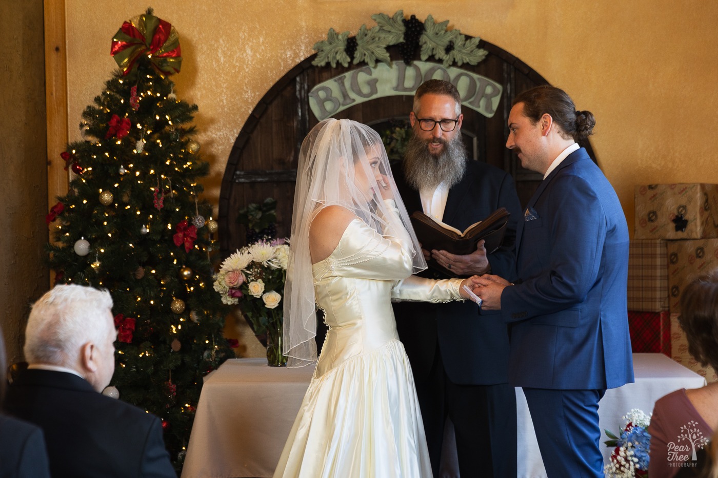 Bride wiping away a tear as her groom holds her hand and smiles while the pastor is talking to them
