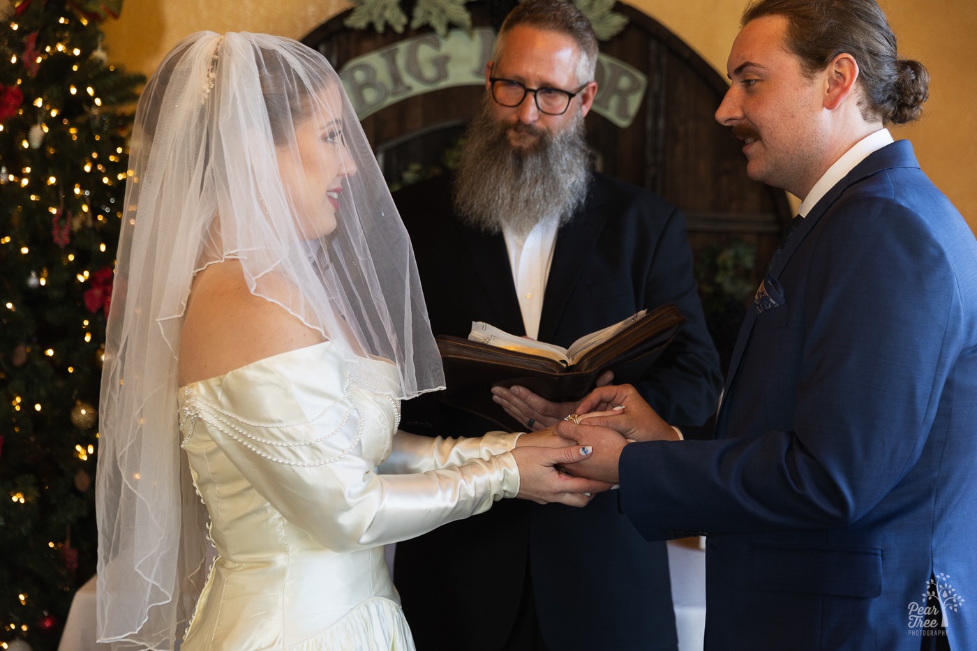Groom slipping a wedding band on his new wife's finger with a Christmas tree and pastor in the background