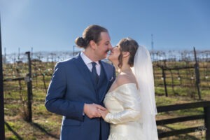 Bride and groom holding hands and smiling as they pull in close for a kiss with a vineyard sparkling under the sun in the background