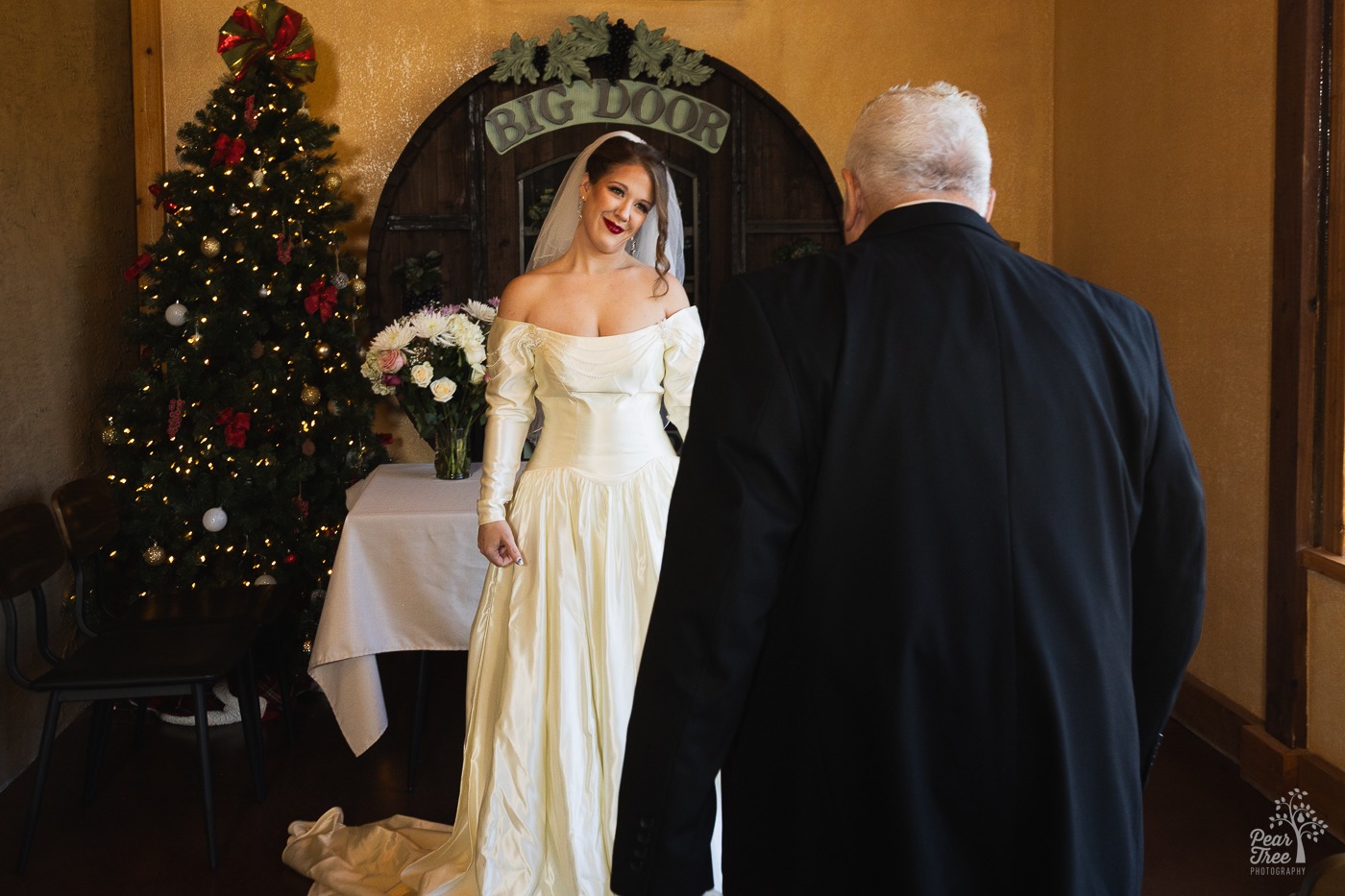 Bride smiling at her dad as he sees her in a wedding dress for the first time. There is a Christmas tree and a Big Door Vineyard sign behind her.