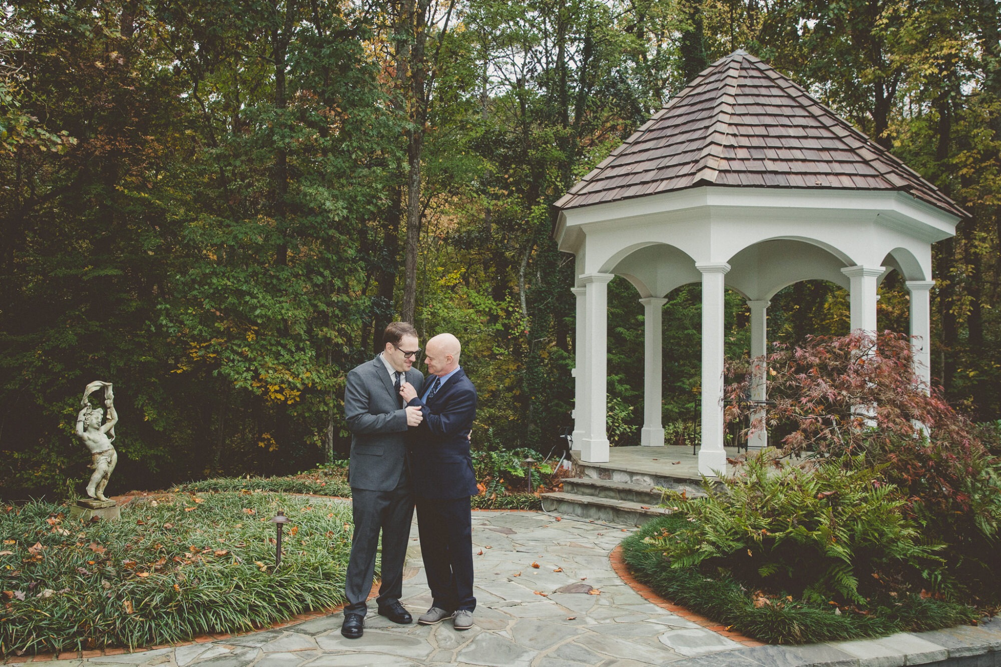 Two grooms in suits holding each other close with a gazebo and statue in the background filled with woods and fall leaves
