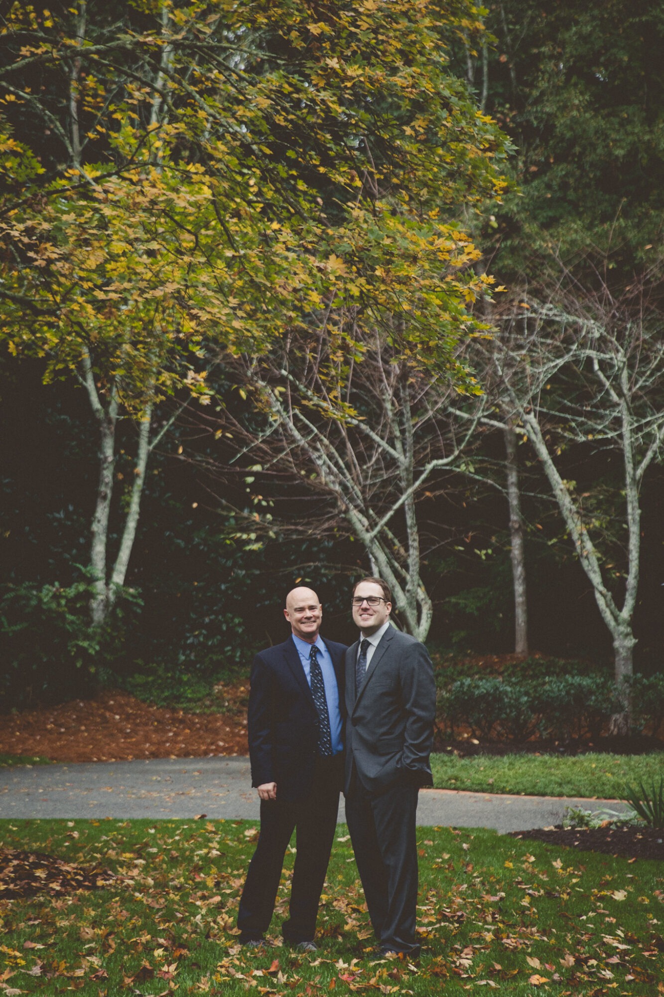 Two grooms in suits smiling at the camera with fall leaves on trees in the background after their backyard Atlanta wedding ceremony