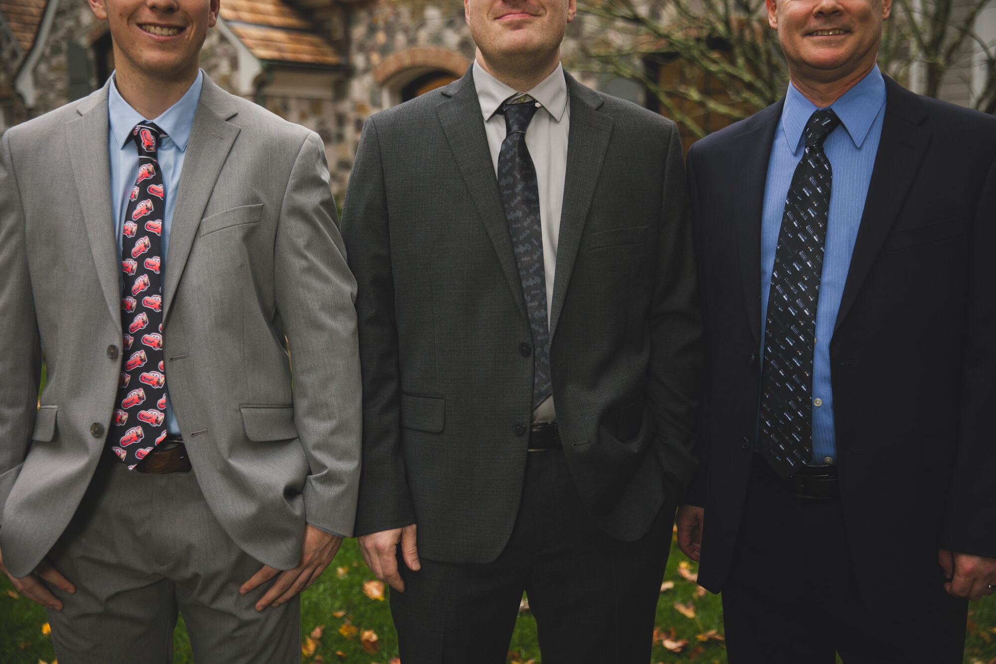 Two grooms and their nephew and wedding officiant smiling in their suits and ties sporting symbols for Batman, Star Wars, and Cars Lightning McQueen