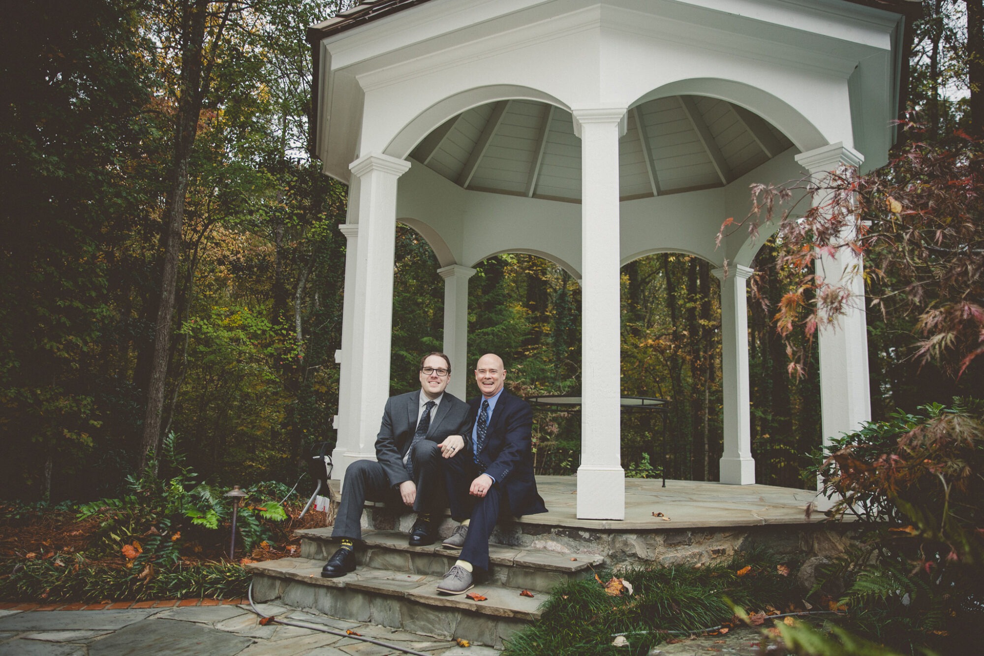 Two grooms in suits sitting on gazebo steps and laughing