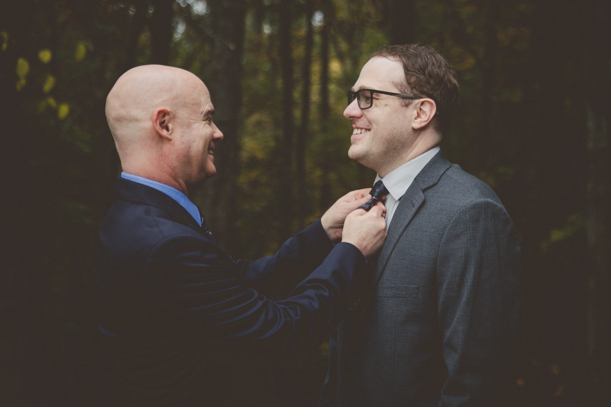 One groom in a navy blue suit straightening the tie of his husband-to-be who is smiling and wearing a grey suit