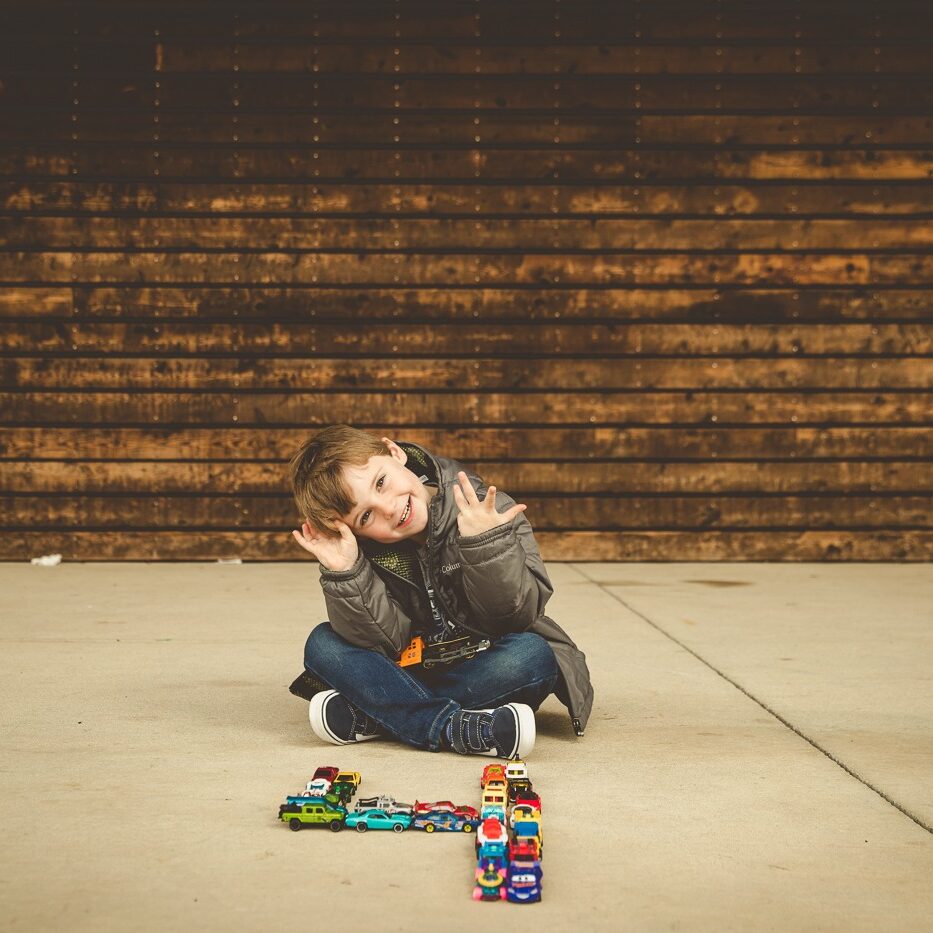 Four year old boy sitting criss cross applesauce behind his toy cars and trains making the number four.