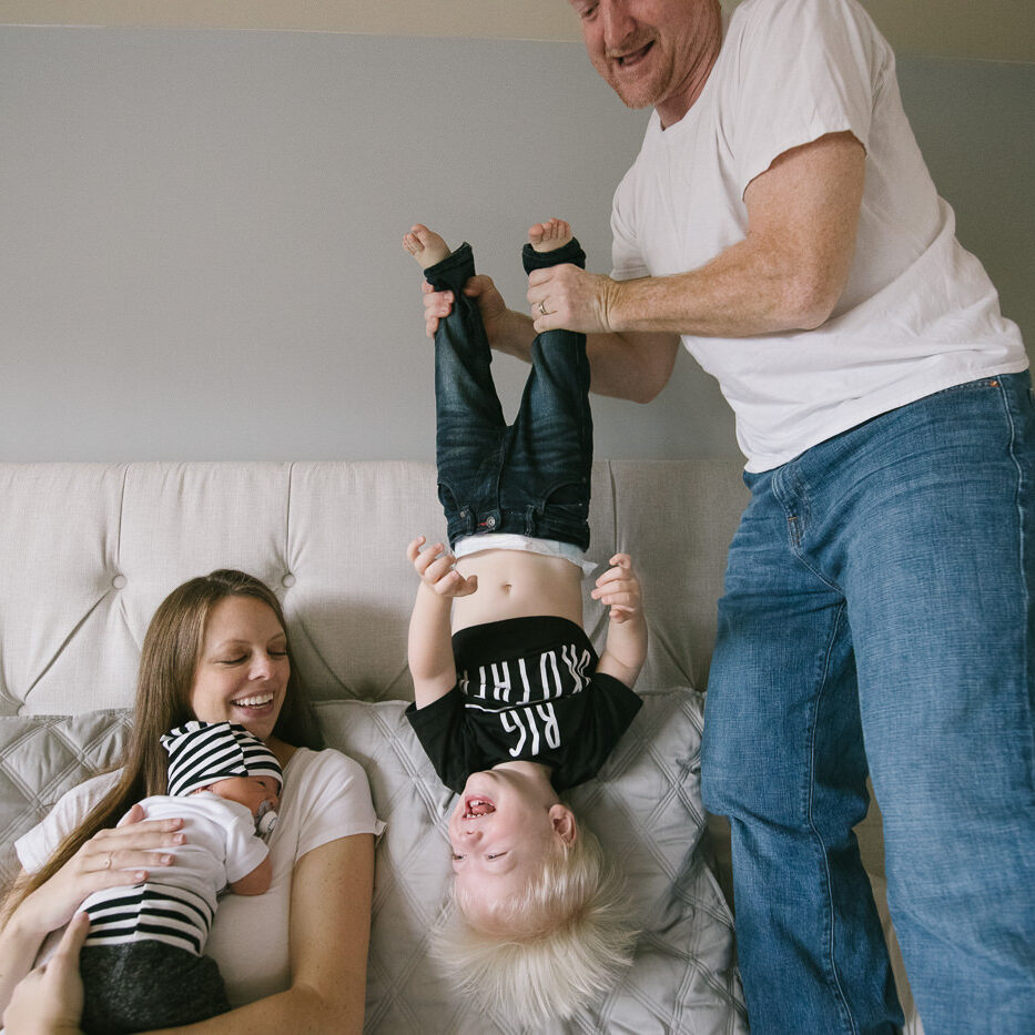 Mom holding newborn son on bed while Dad stands and holds their toddler upside down