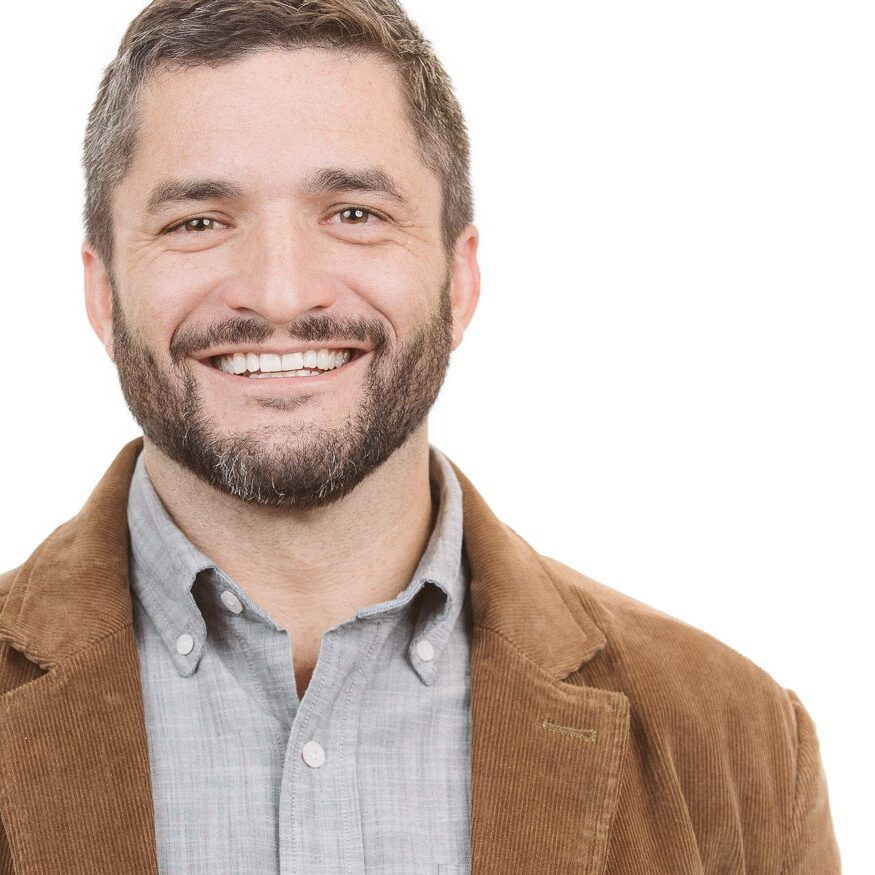 Headshot of a smiling white male with mustache and beard wearing a brown corduroy blazer and plaid button down shirt