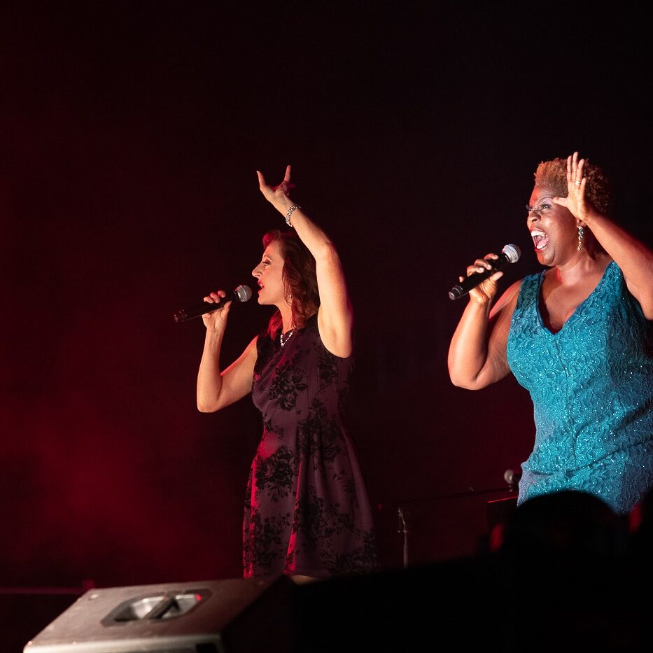 Broadway stars Rita Harvey and Capathia Jenkins performing live with microphones to mouths and left hands raised in the air while they belt out a song wearing stunning dresses
