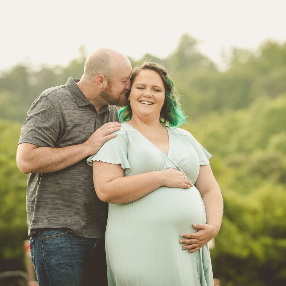 Pregnant mom holding her belly in a green dress. Her husband has her hand on her shoulder and is pushing his nose into her ear