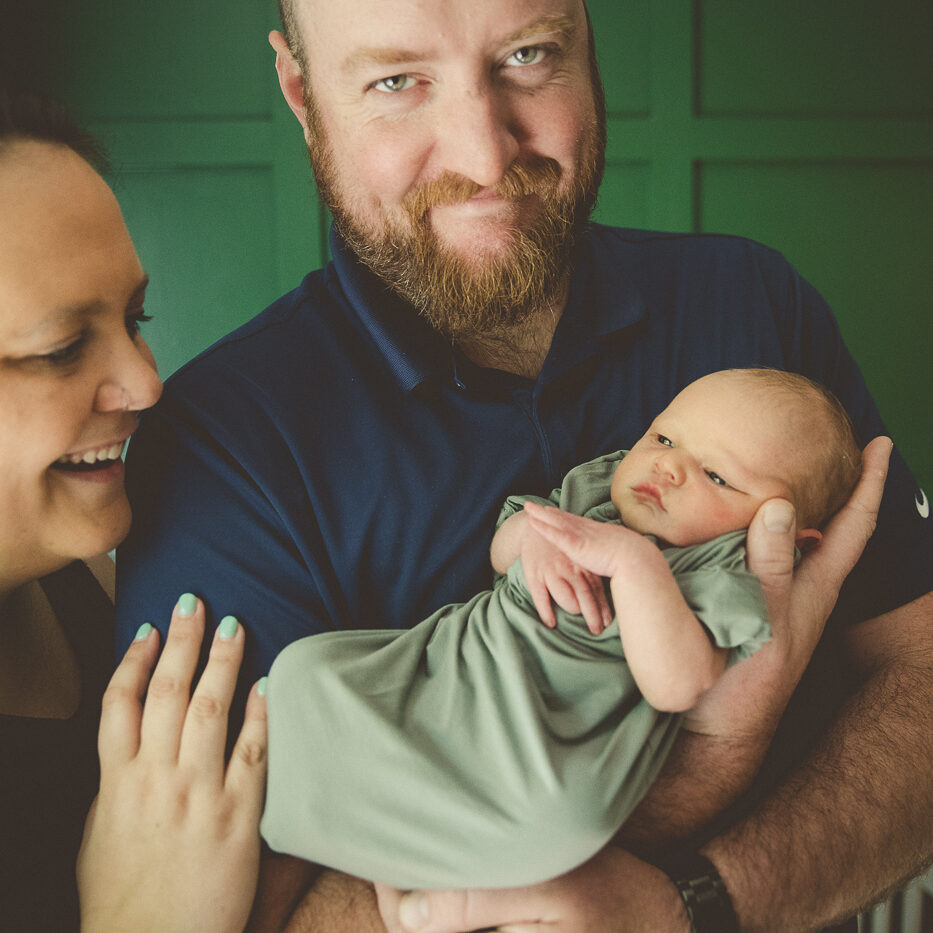 Proud dad holding his newborn son with mom smiling and her hand on his arm during their newborn photography session