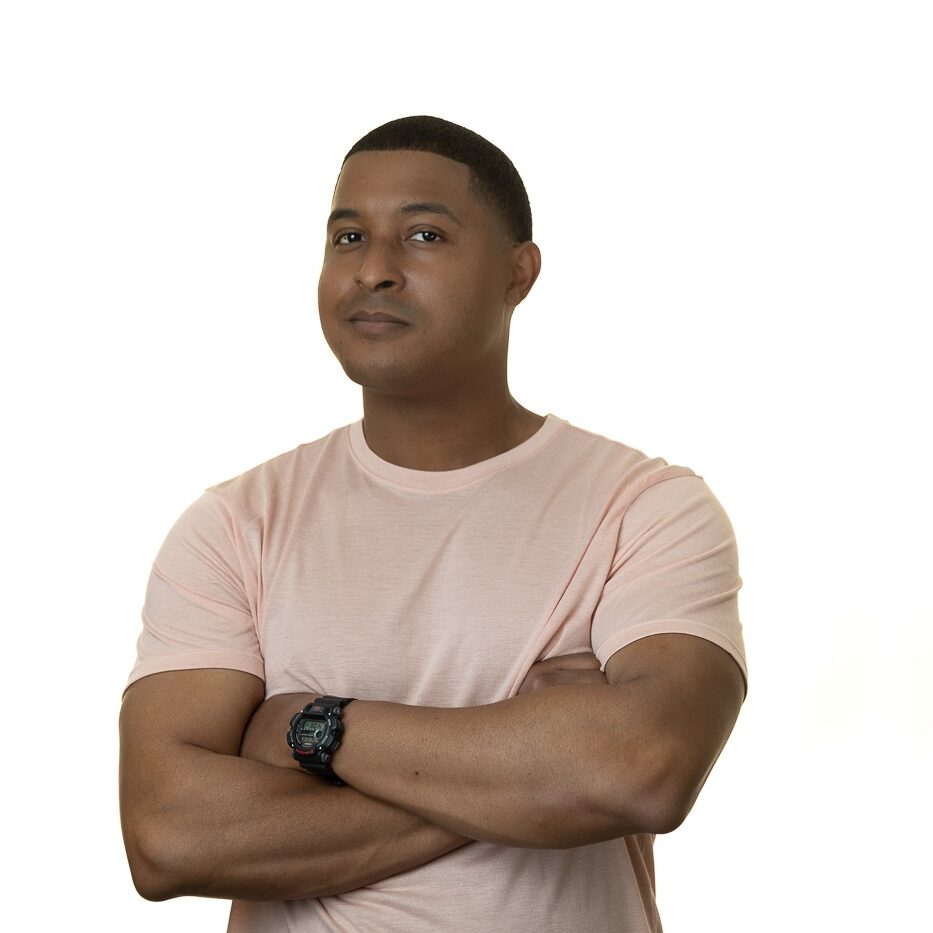 Professional headshot of an attractive African American male. He's smirking with his arms crossed while wearing a black watch, pink shirt and standing in front of a white background.