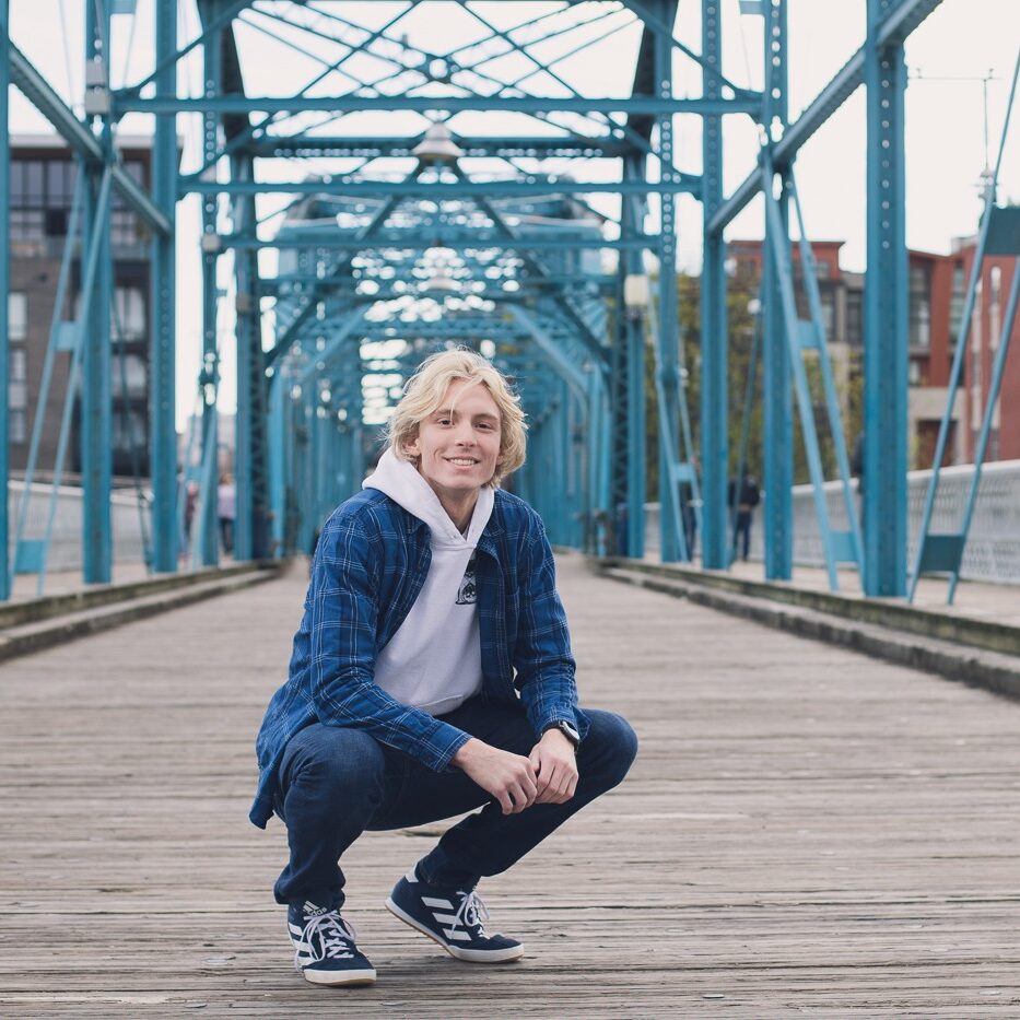 High school senior boy wearing jeans and squatting on bridge Chattanooga bridge