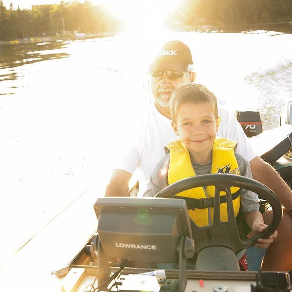 Little boy sitting in his Grandpa's lap while driving a boat across a lake at sunset.