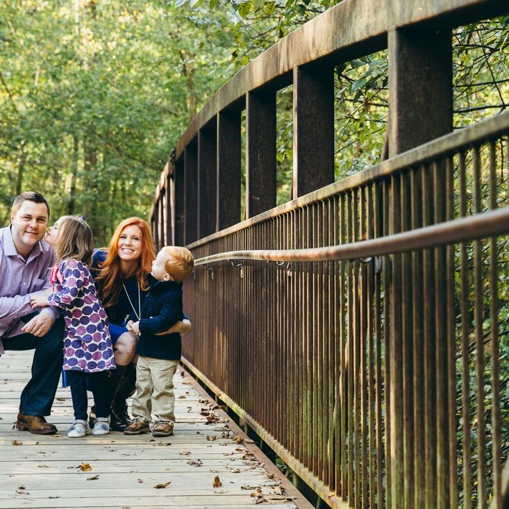 Family of four with two littles smilling and smooching on bridge in East Cobb park