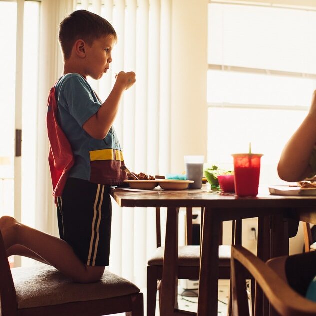 Young boy dressed like Superman eating breakfast at the kitchen table with his little brother